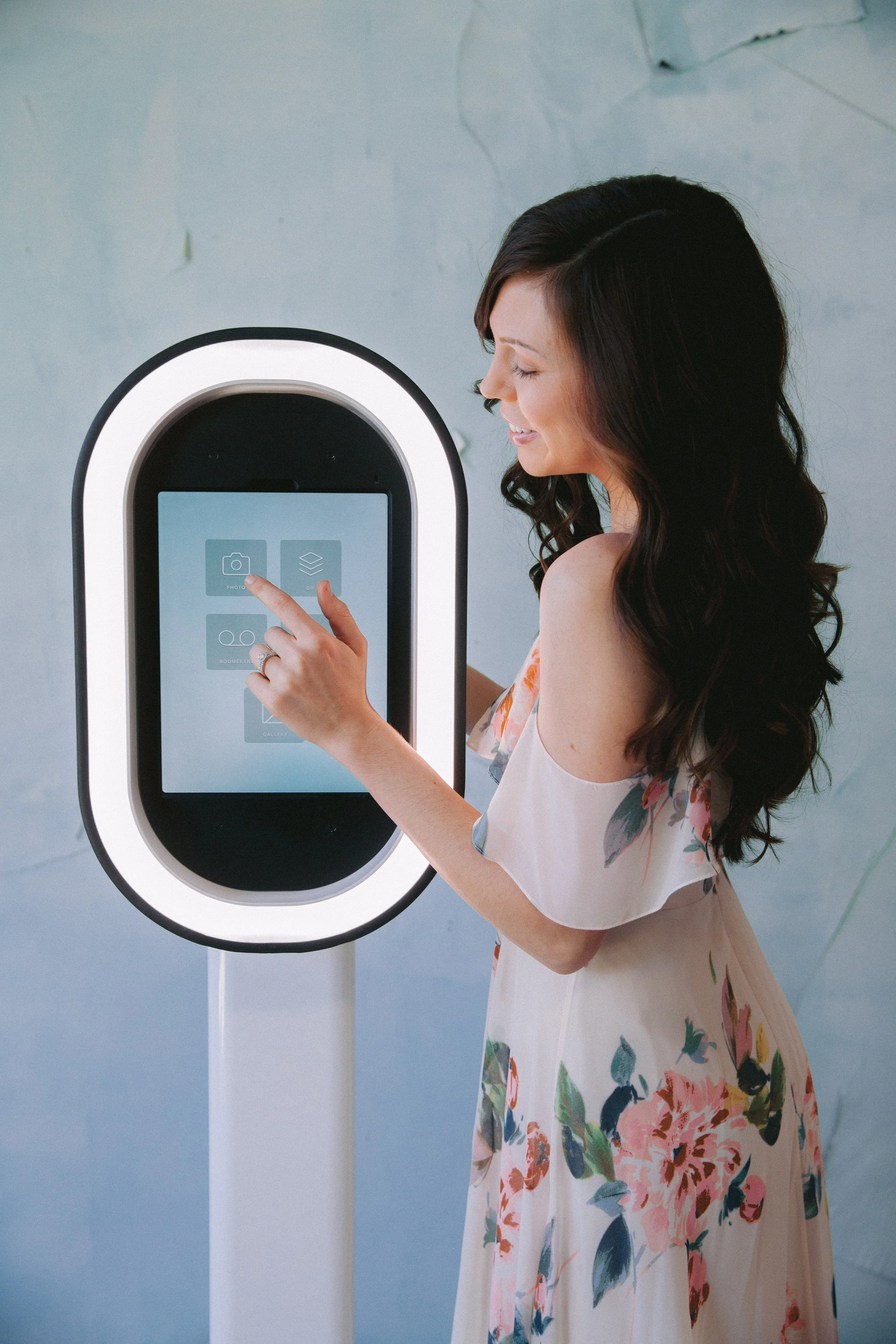 A woman with long, dark, wavy hair in a floral dress using a touchscreen kiosk with a white frame and circular light outline.