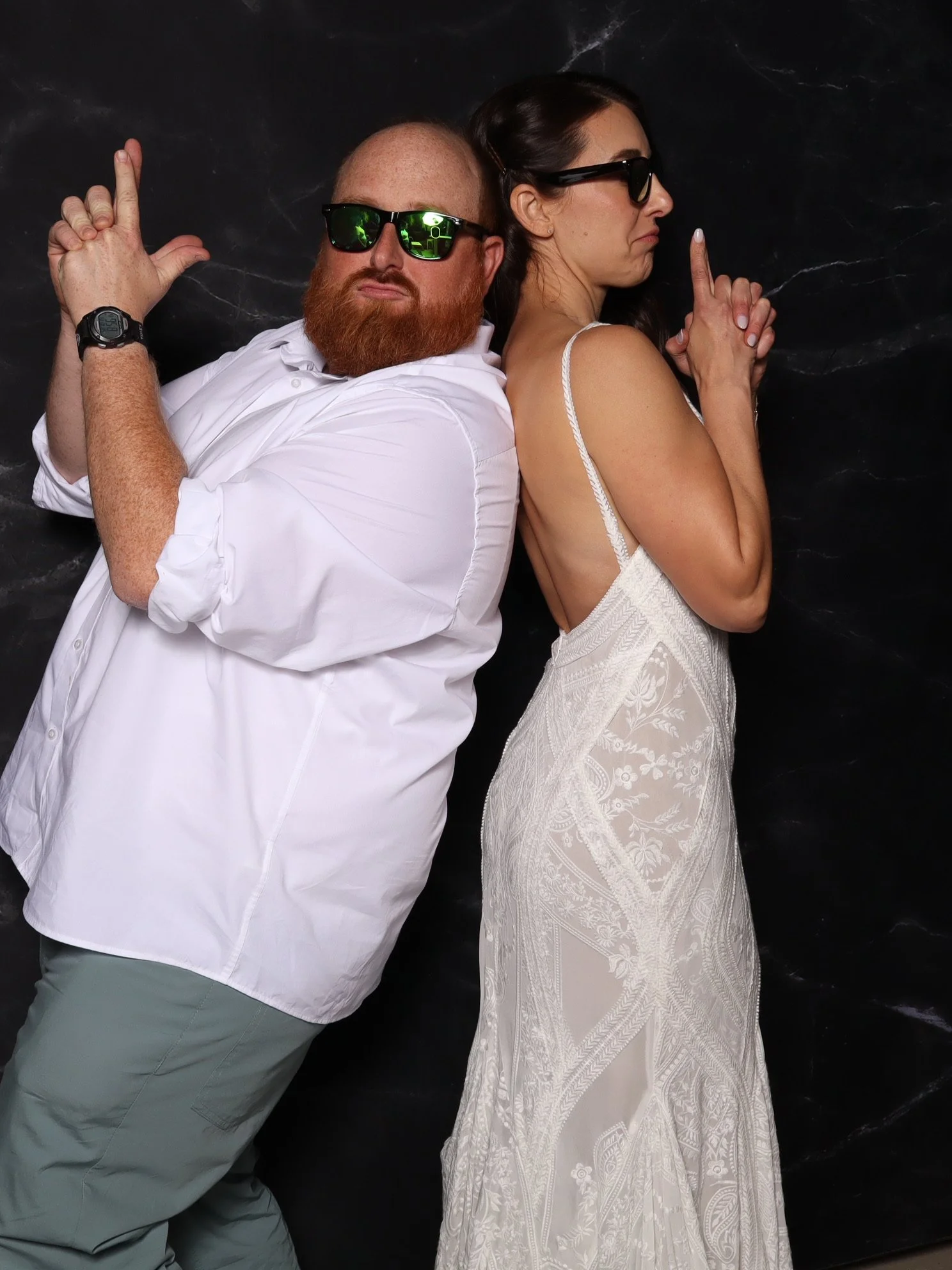 Two people, a man with a beard wearing sunglasses and a white shirt, and a woman with dark hair in sunglasses and a white dress, are sitting back-to-back with arms crossed in a 'finger gun' pose against a dark background.