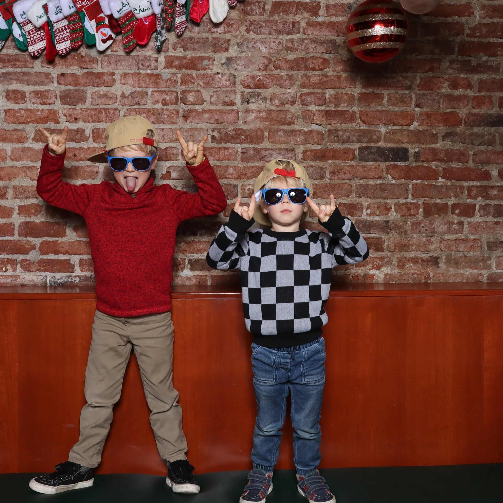 Two young boys wearing sunglasses and caps posing in front of a brick wall decorated with Christmas stockings and a large red ornament.
