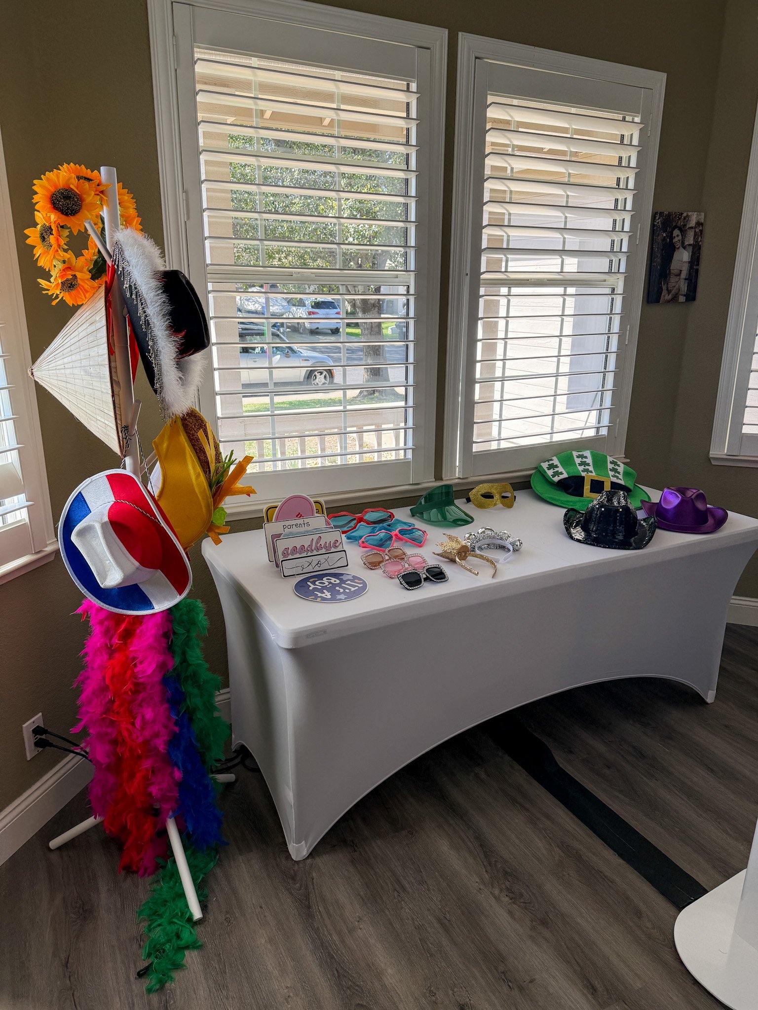 Table with party props including hats, glasses, masks, and a sign that says "goodbye". A colorful feather boa and a small stand with hats and sunflowers are nearby. Behind are two large windows with white blinds, and a photo on the wall.