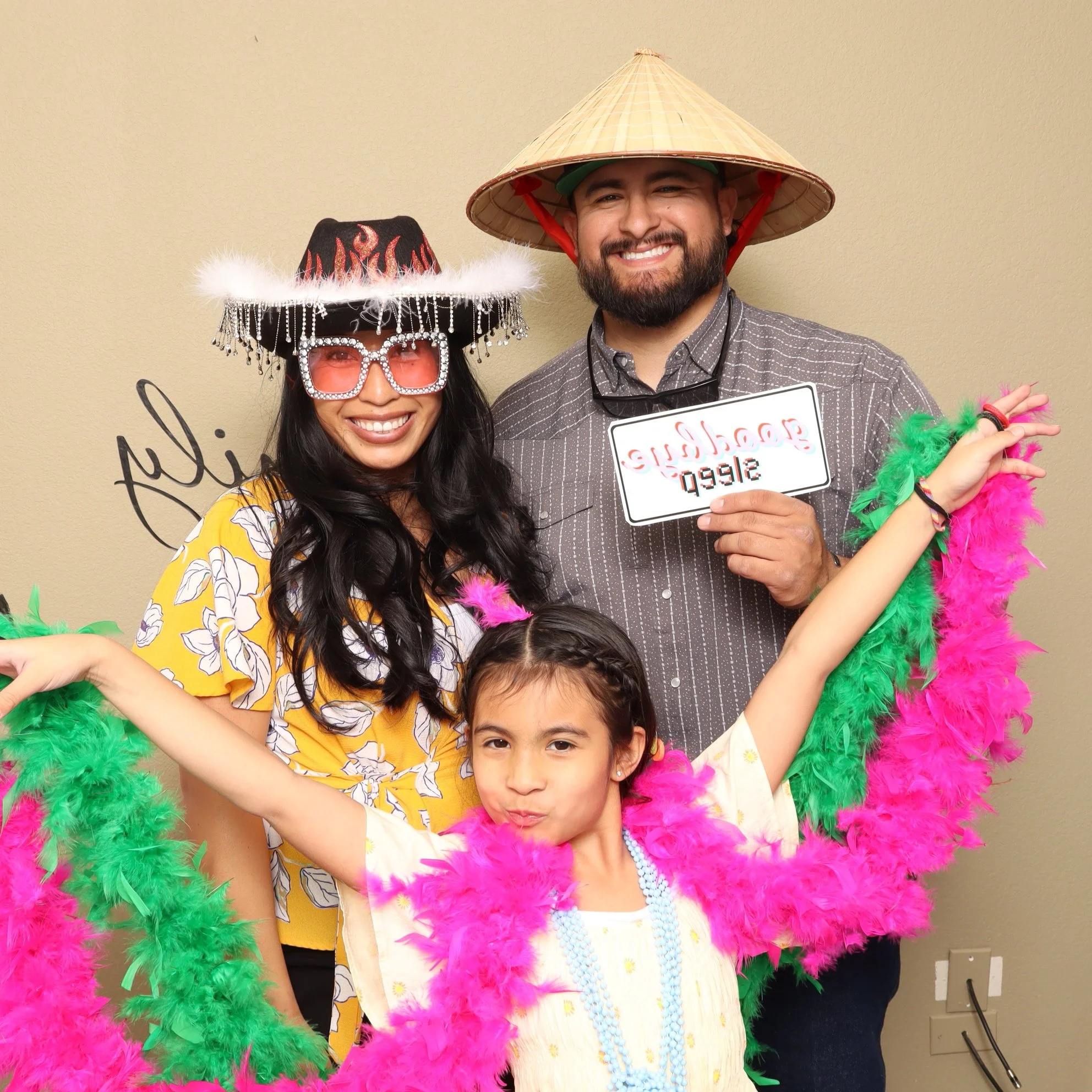 Three people celebrating at a photo booth with colorful props including feather boas, hats, and glasses. The man wears a traditional Asian conical hat; the woman wears a cowboy hat with flames and beaded glasses; the girl has her arms outstretched and is wearing a feather boa and beaded necklace.