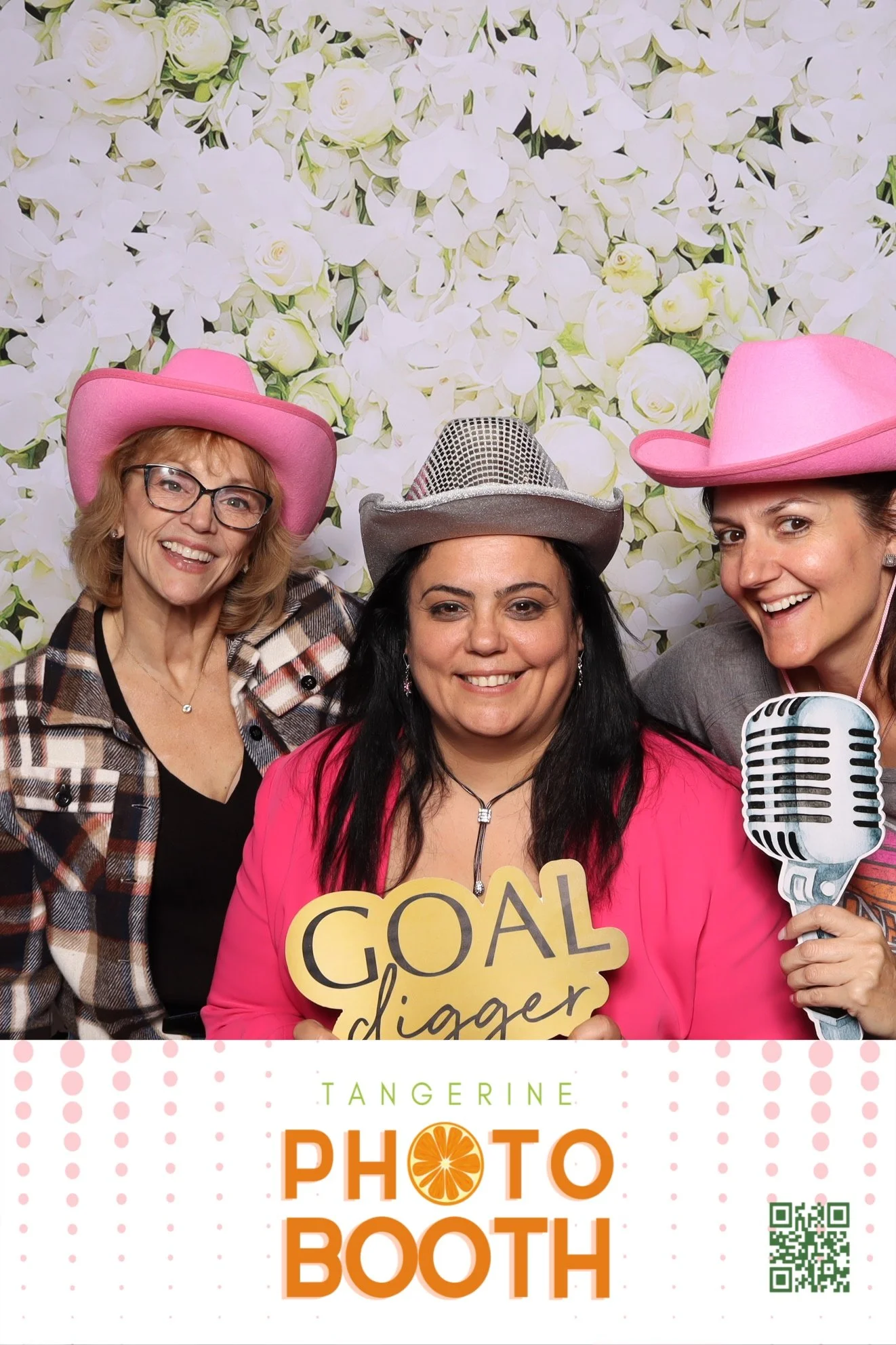 Three women smiling in a photo booth with floral backdrop, wearing pink and silver cowboy hats, one holding a sign that says 'Goal Digger', and another holding a microphone cutout, at the 'Tangerine Photo Booth' event.
