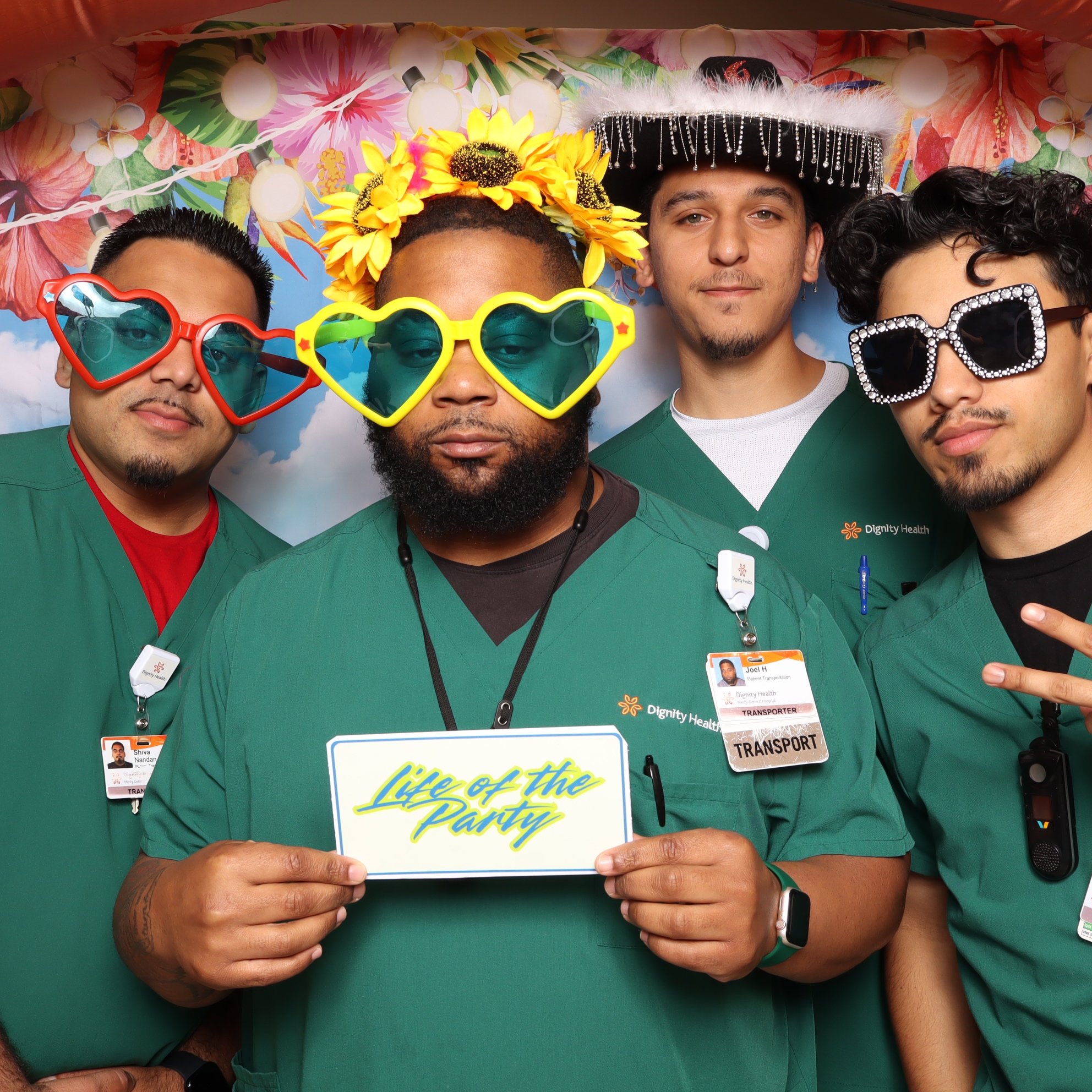 Four healthcare workers wearing green scrubs and large, colorful sunglasses, standing in front of a floral background with string lights, some with playful hats, one holding a sign that says 'Life of the Party'.