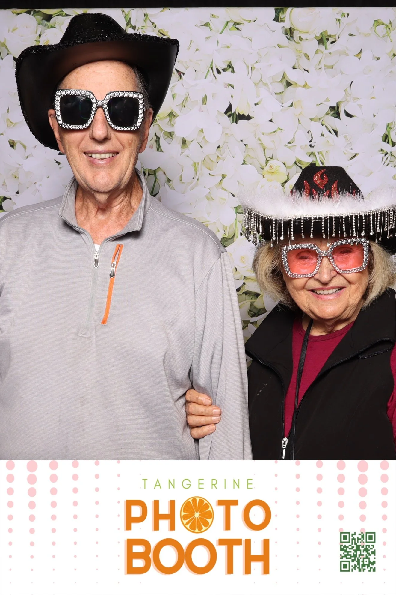 A smiling older man and woman wearing fun hats and oversized sunglasses in front of a white floral backdrop at a photo booth. The man has a black cowboy hat and large sunglasses decorated with rhinestones, while the woman has a black hat with red flames, white feathers, and rhinestones, and pink sunglasses with rhinestones.