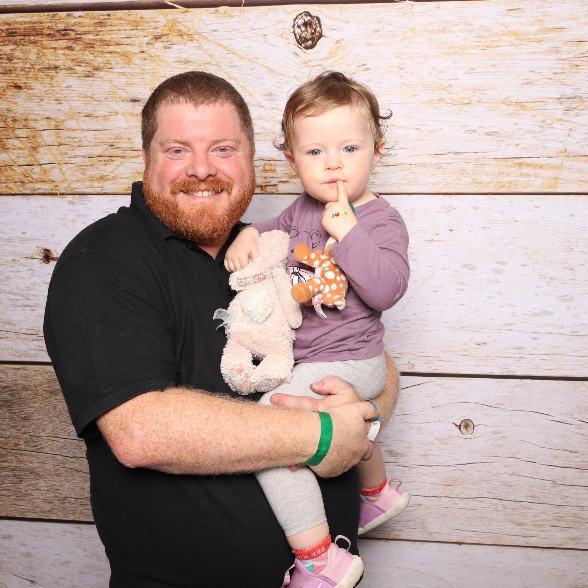 A man with a red beard and wearing a black shirt holding a young girl with curly blonde hair, wearing a purple shirt and gray pants, in front of a wooden background. The girl is holding stuffed animals and has her finger to her lips.