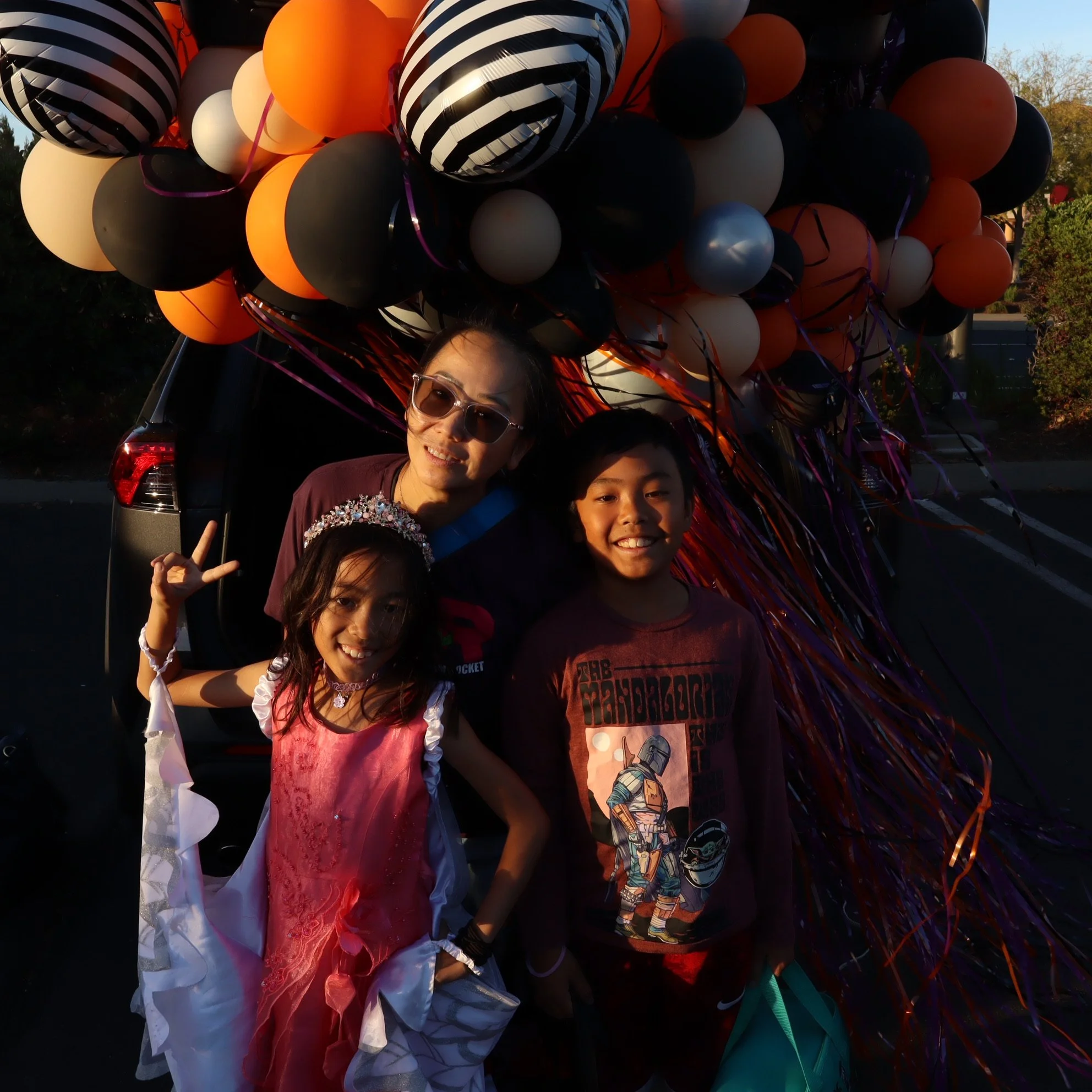 A woman and two children standing in front of a car decorated with numerous black, orange, white, and striped balloons, celebrating an outdoor event during sunset.