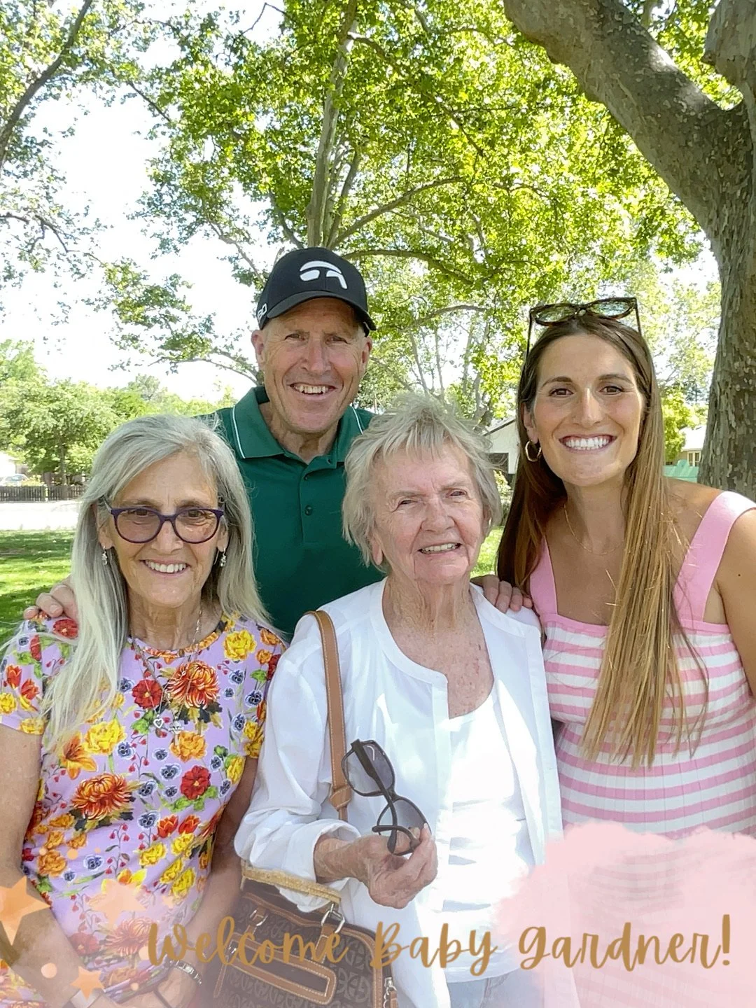 A group of five people, three women and two men, smiling outdoors under a large green tree, with a caption that says "Welcome Baby Gardner!"