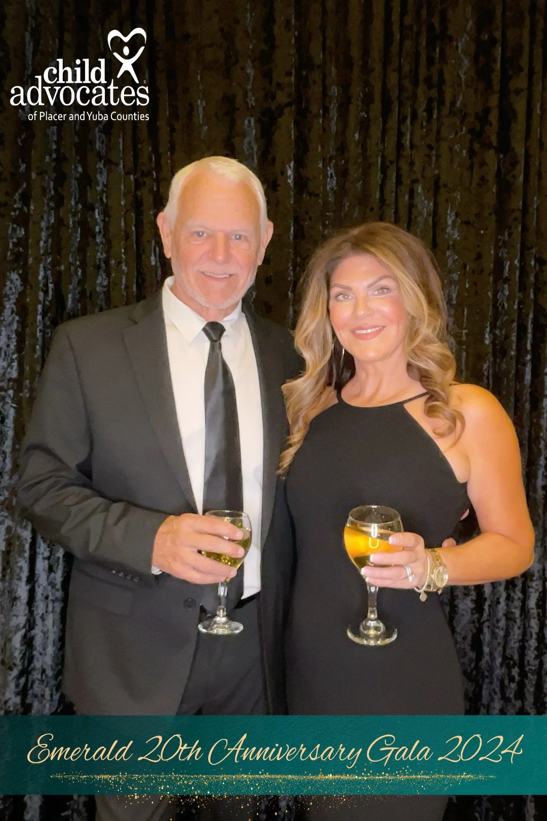 A man in a tuxedo and a woman in a black dress holding glasses of wine at an event, standing in front of a black curtain, celebrating the Emerald 20th Anniversary Gala 2024, organized by child advocates of Placer and Yuba Counties.