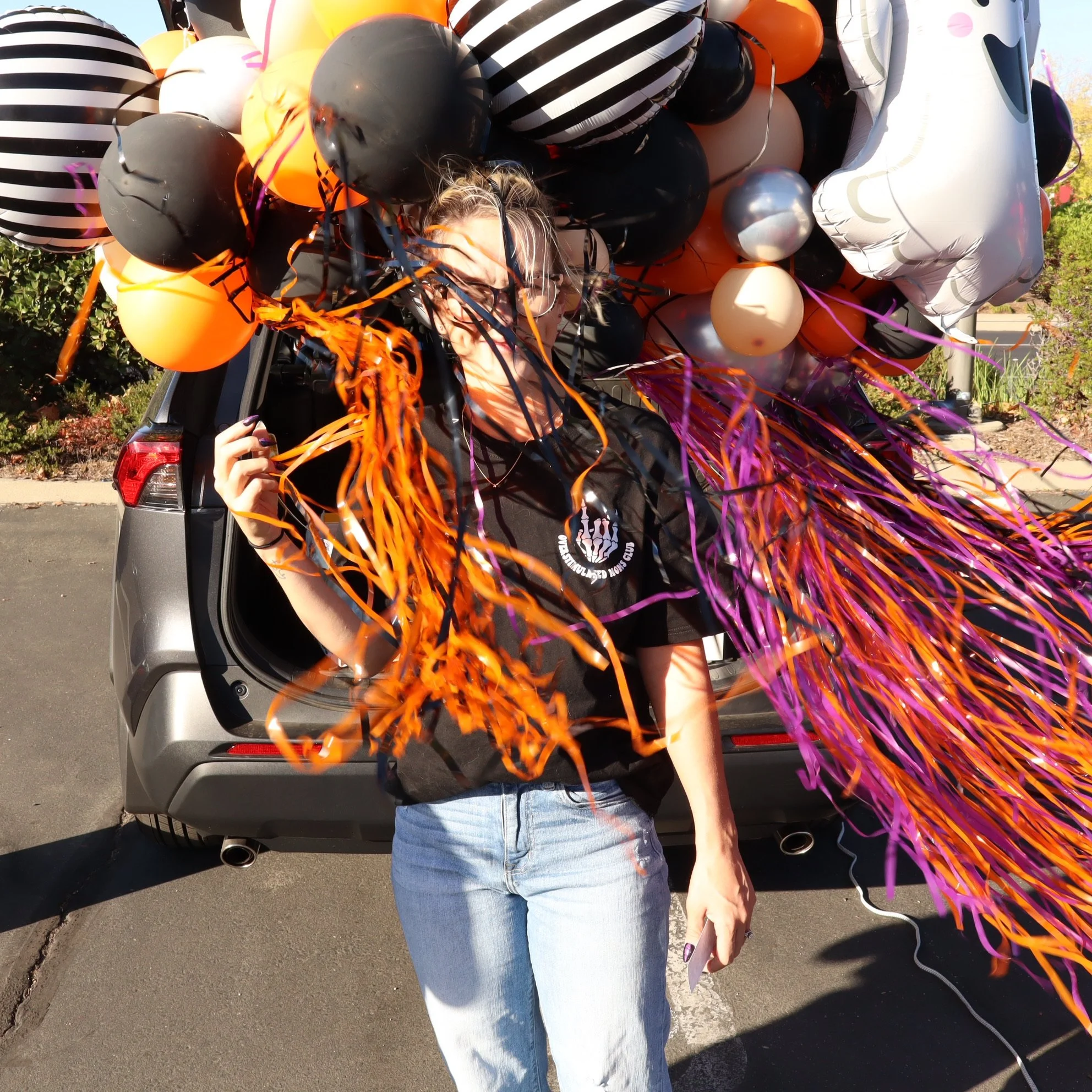A woman standing in front of an open car trunk decorated with black, orange, purple, and silver balloons and streamers, celebrating Halloween or a festive event.