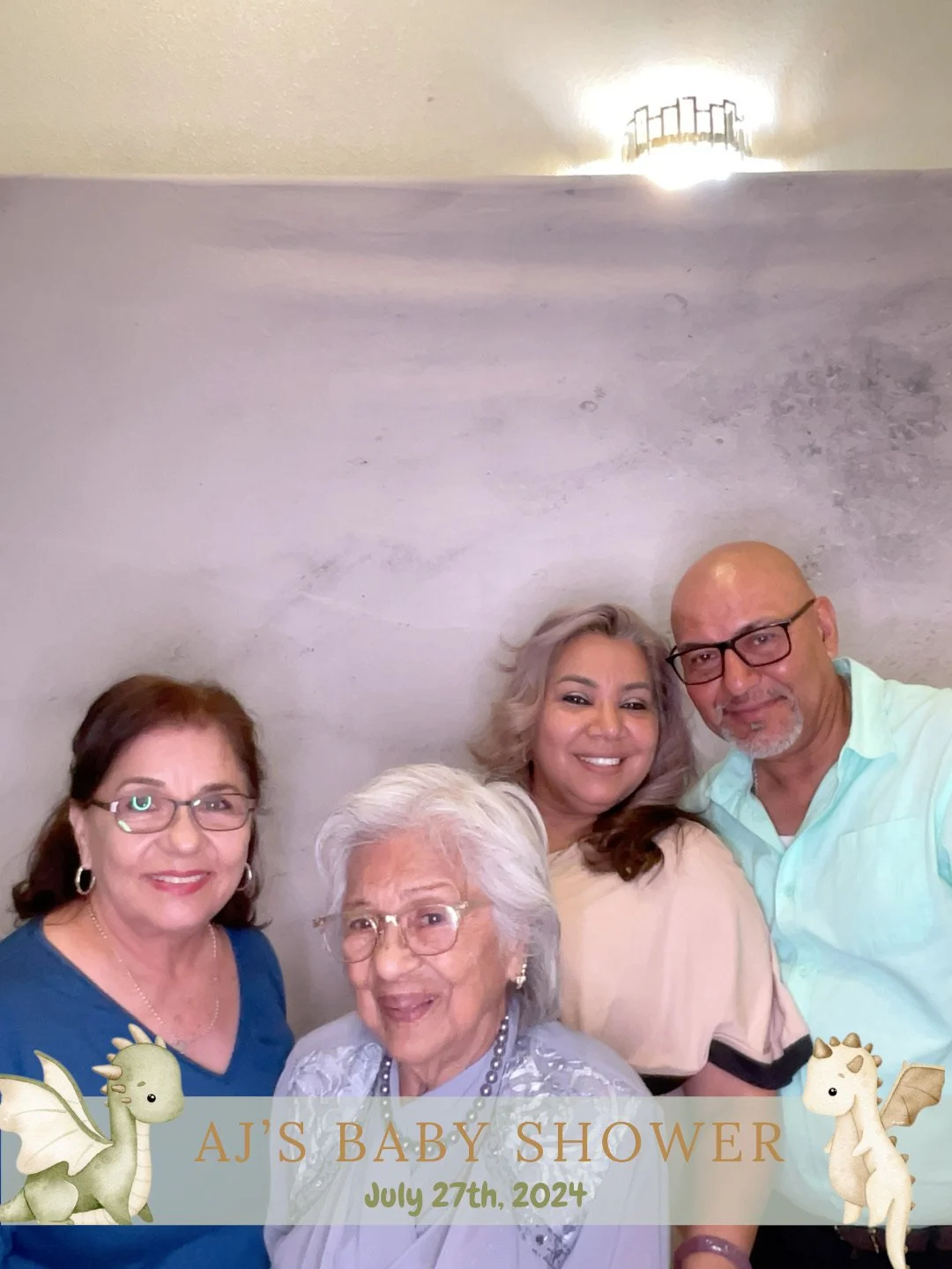 Four women posing together at a baby shower with a banner that reads "AJ'S BABY SHOWER July 27th, 2024" featuring cute dragon illustrations.