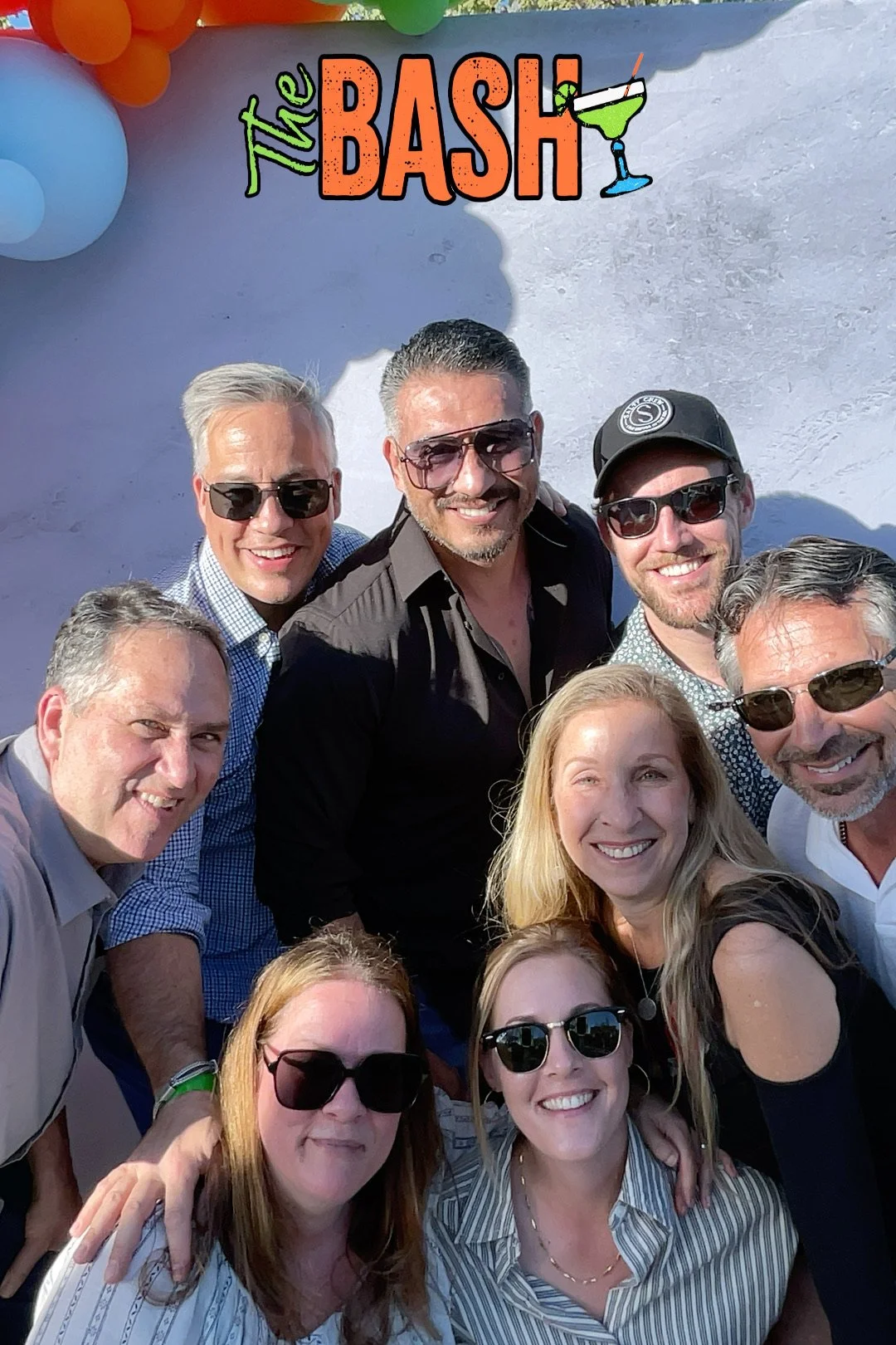 A group of nine adults smiling for a selfie at a party called 'The Bash,' with balloons in the background and the event's colorful sign at the top.