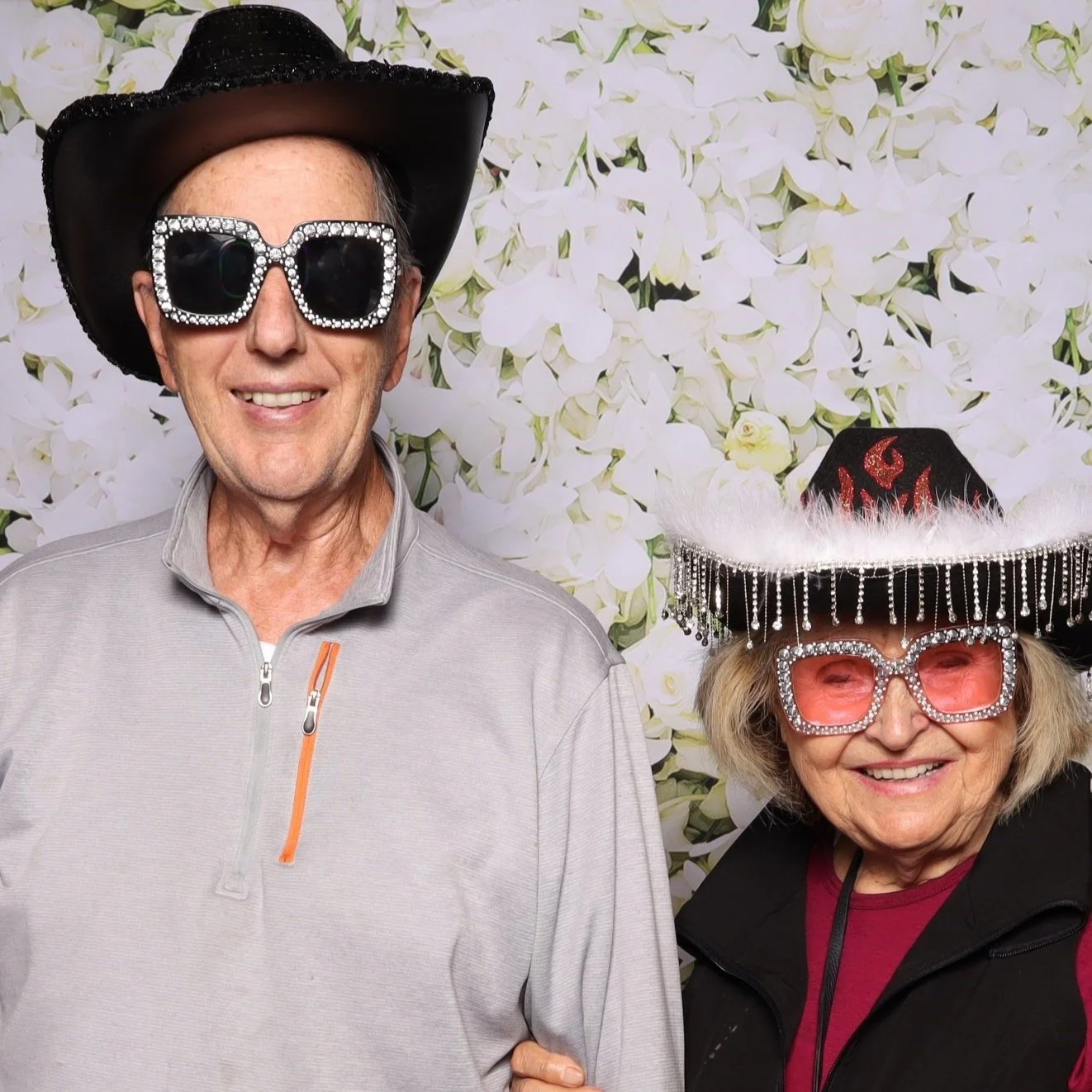 Two older women wearing large, sparkly sunglasses and decorated hats posing together in front of a floral background.
