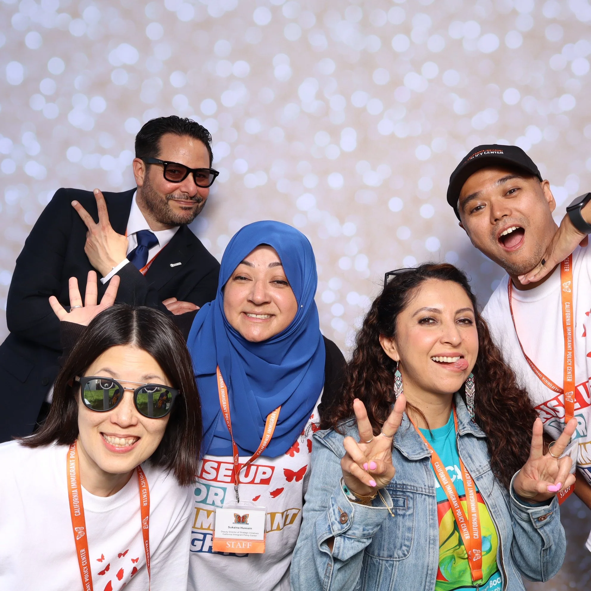 Group of five diverse people at an event, smiling and making hand gestures, with a sparkly background.