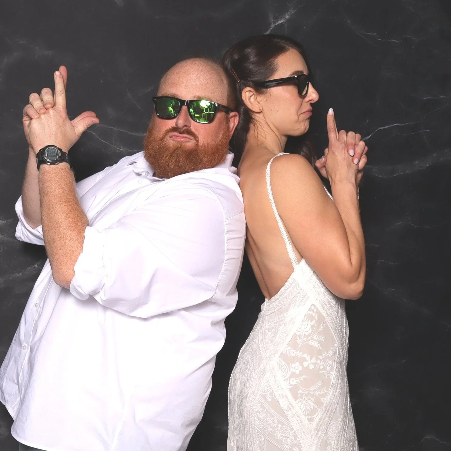 A man and a woman back-to-back with sunglasses, posing in a playful manner. The man has a beard, bald head, white shirt, and a watch. The woman has dark hair, a white dress with thin straps, and is making a gesture with her finger.