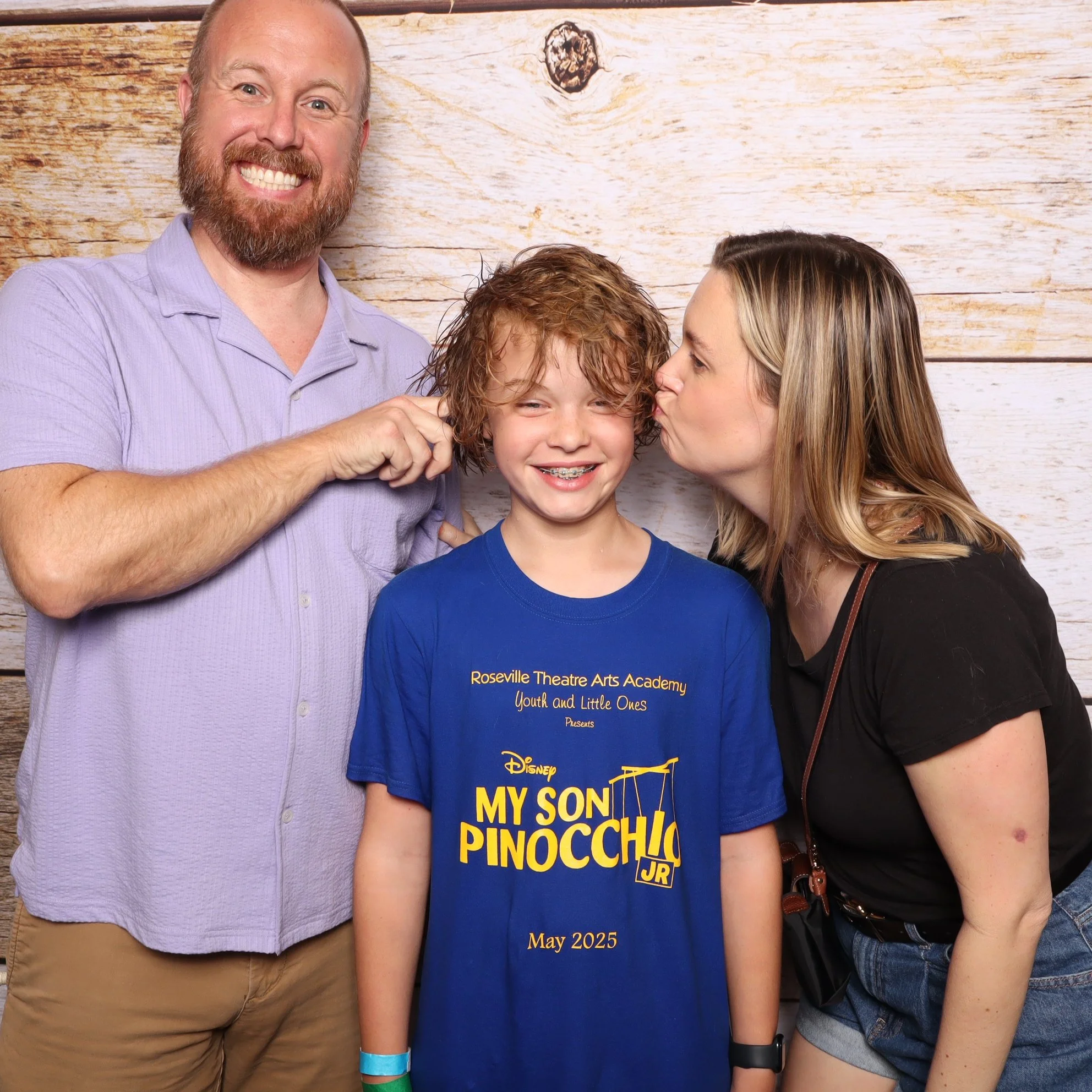 A smiling older man with a beard tugging on the ear of a smiley boy with curly red hair, who is wearing a blue 'My Son Pinocchio Jr.' T-shirt. A woman with blonde hair is kissing the boy on the cheek. They are standing in front of a wooden wall.