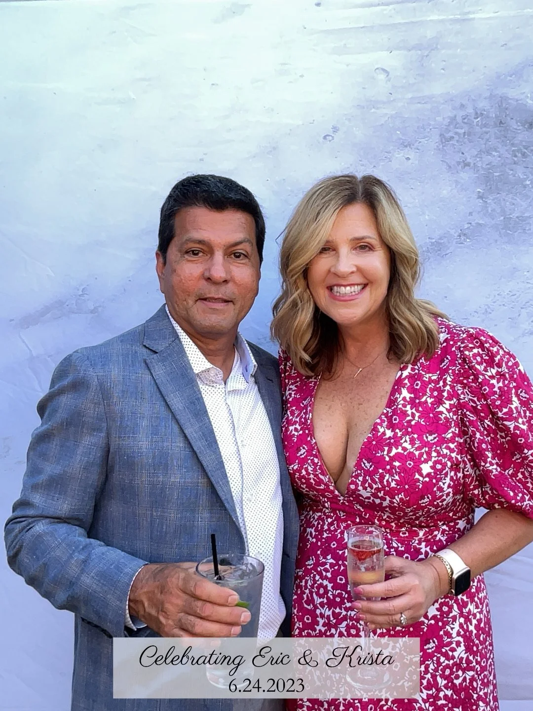 A man in a gray suit with a white shirt holding a drink, standing next to a woman in a pink floral dress also holding a drink, both smiling, at a celebration event with a white backdrop. There is a caption that reads 'Celebrating Eric & Krista 6.24.2023'.