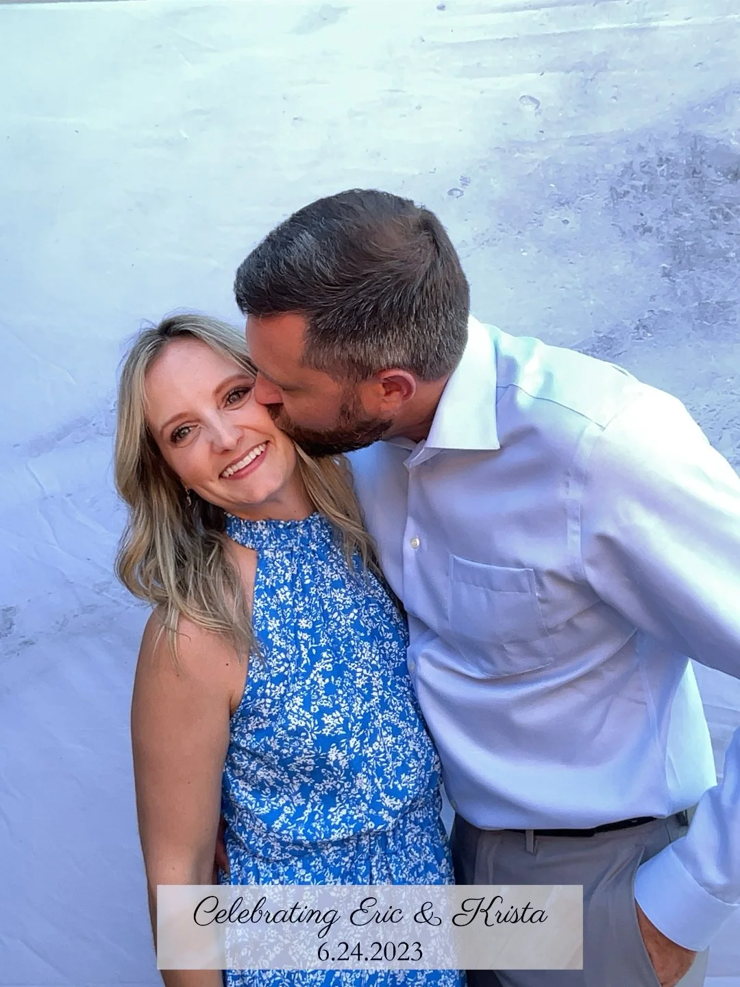 A couple celebrating Eric and Krista on June 24, 2023, with the man kissing the woman on the cheek. The woman is smiling and wearing a blue dress, and the man is dressed in a white shirt. They are standing in front of a light-colored background.
