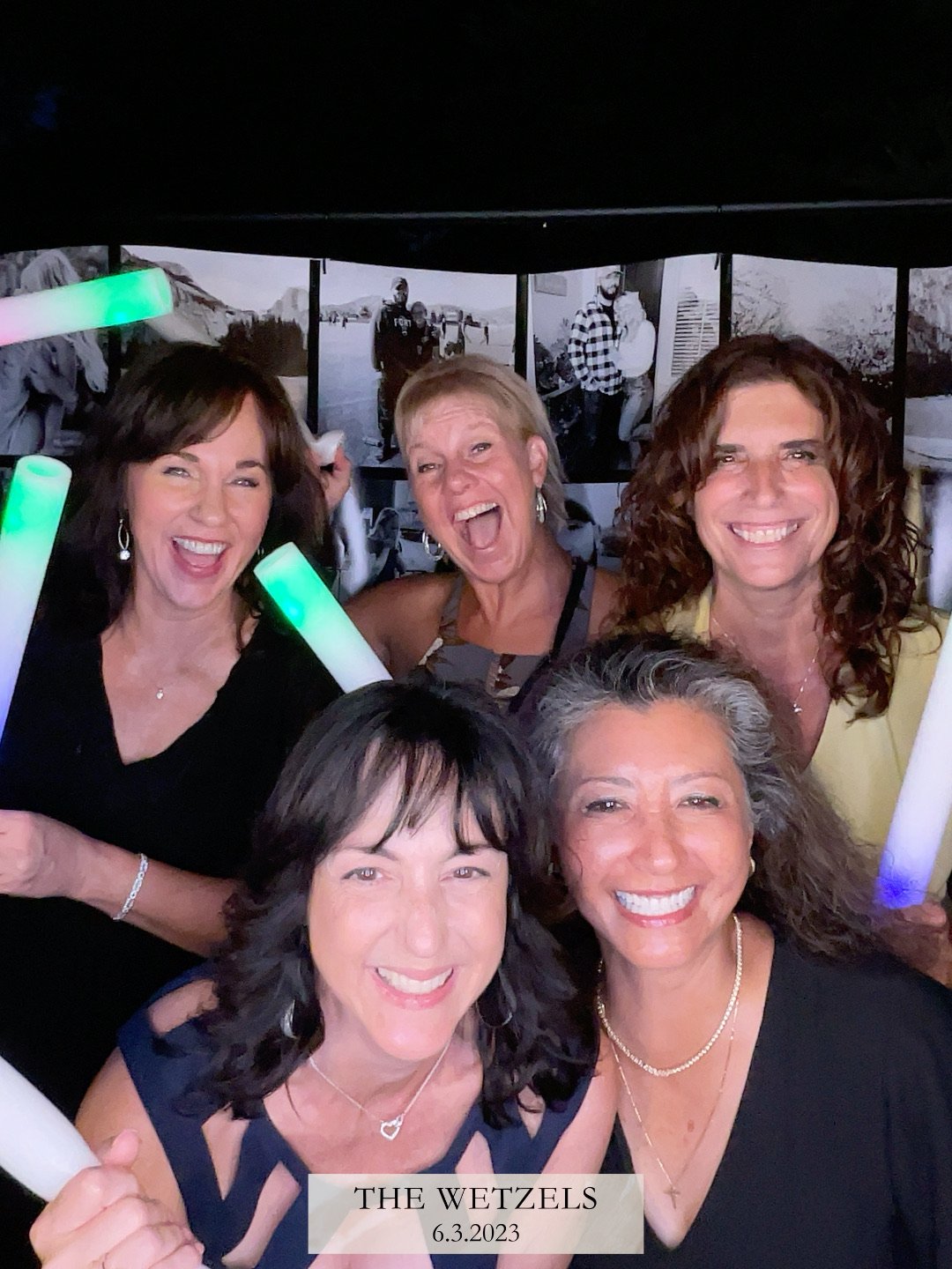 Group of five women at a celebration event, smiling and holding glow sticks, with photos on a black and white background wall. A banner at the bottom reads "The Wetzels 6.3.2023."