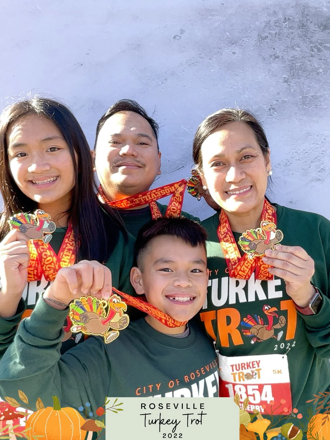 A group of four smiling people—two women, one man, and one young boy—holding Turkey Trot medals in front of a plain, light-colored wall at a 2022 Turkey Trot event in Roseville, dressed in green and orange athletic shirts with the event logo.