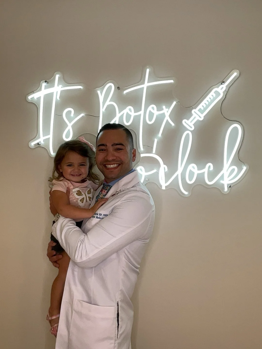 A man in a white medical coat holding a smiling young girl in front of a neon sign that says 'It's Botox o'clock' with a syringe illustration.