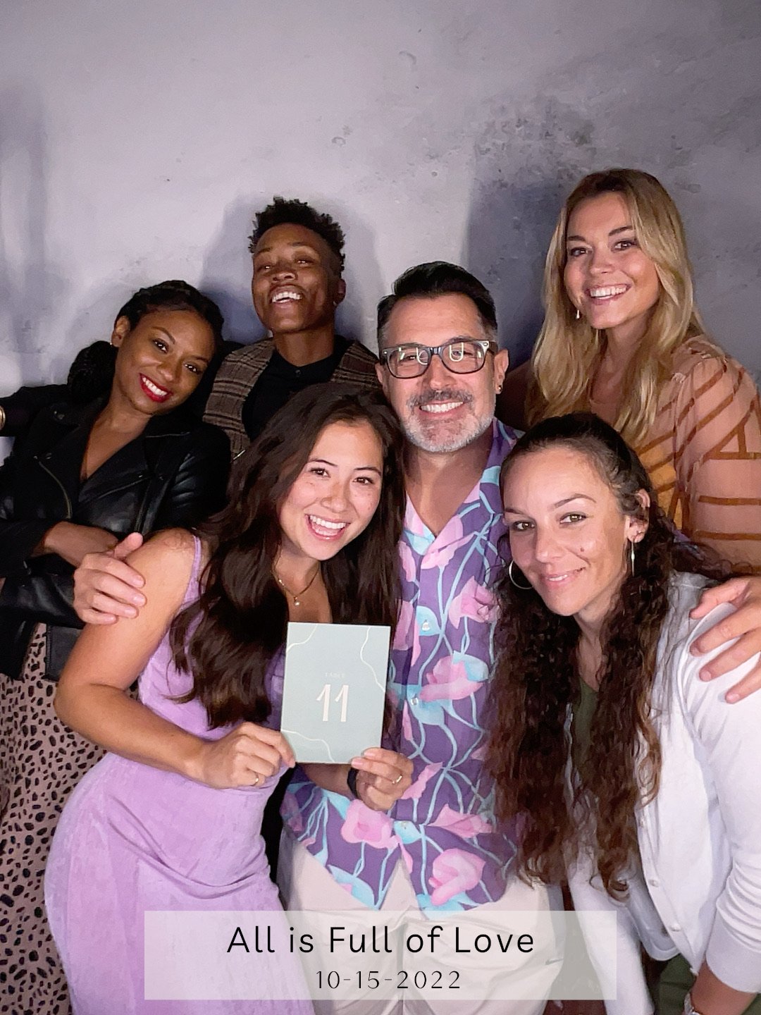 Group of seven diverse people smiling and posing for a photo, with a woman holding a sign with the number 11, at an event titled 'All is Full of Love' on October 15, 2022.