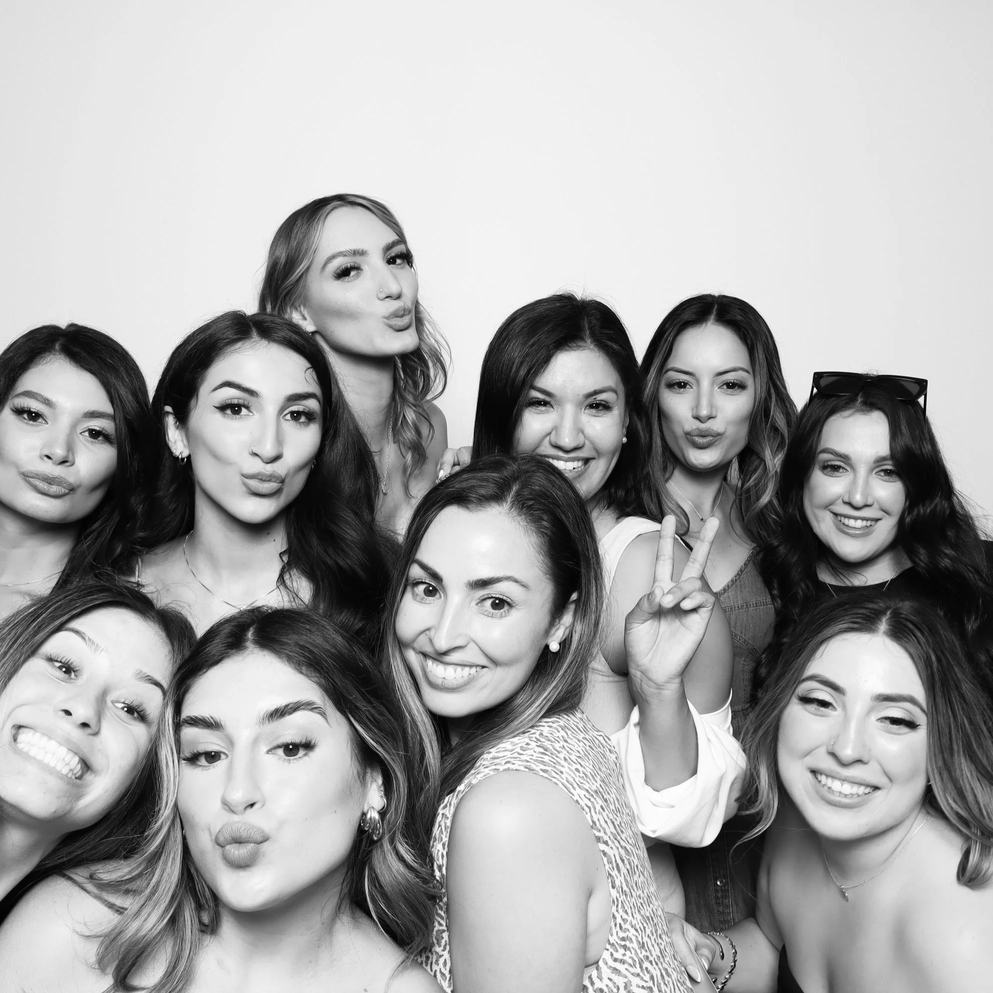 Black and white photo of nine young women taking a group selfie, with some making kissy faces, smiling, or flashing peace signs, against a plain background.