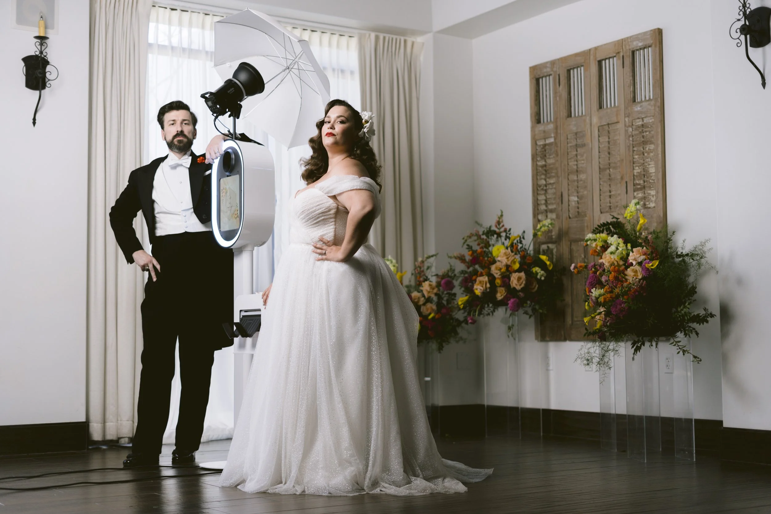 A bride and groom at a wedding photoshoot, with the bride in a white wedding dress and the groom in a tuxedo, standing near photo equipment inside a room decorated with flowers and wall sconces.