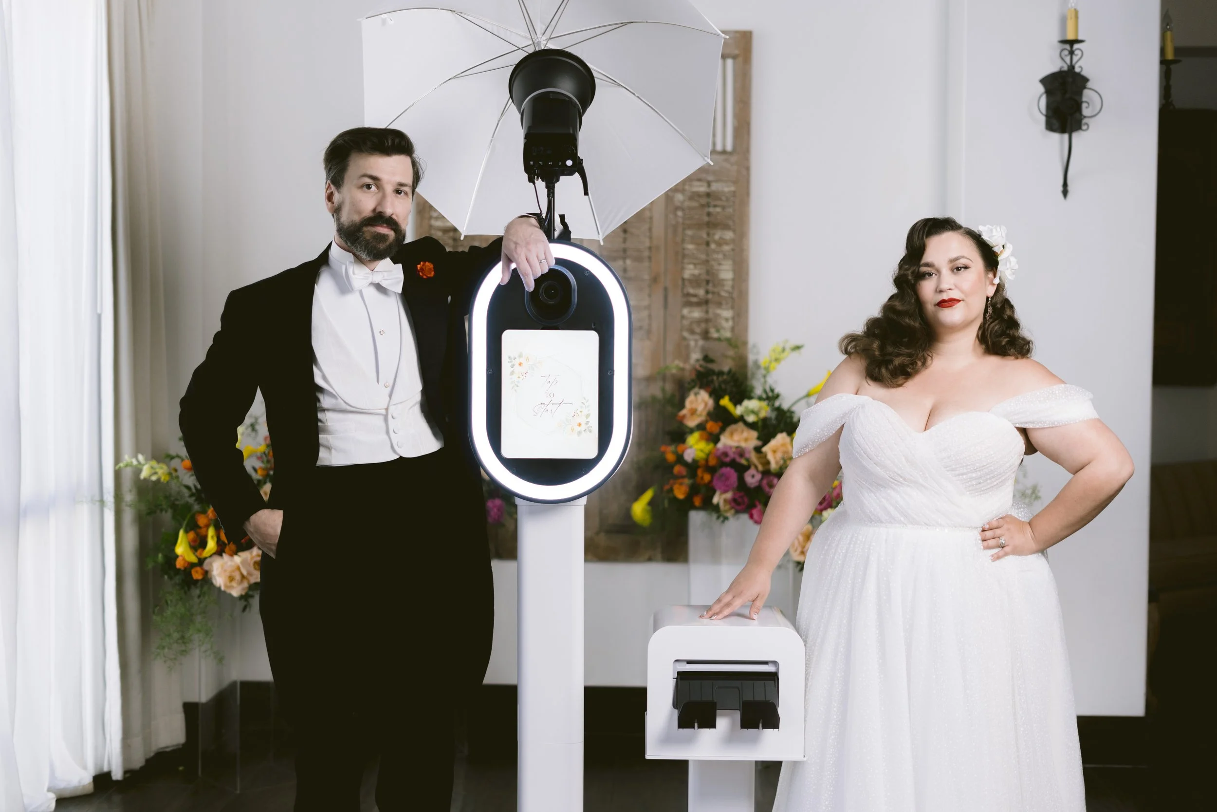 A man in a tuxedo and a woman in a white wedding dress pose next to a photo booth with a ring light and umbrella setup. The setting includes floral decorations and a brick wall backdrop.