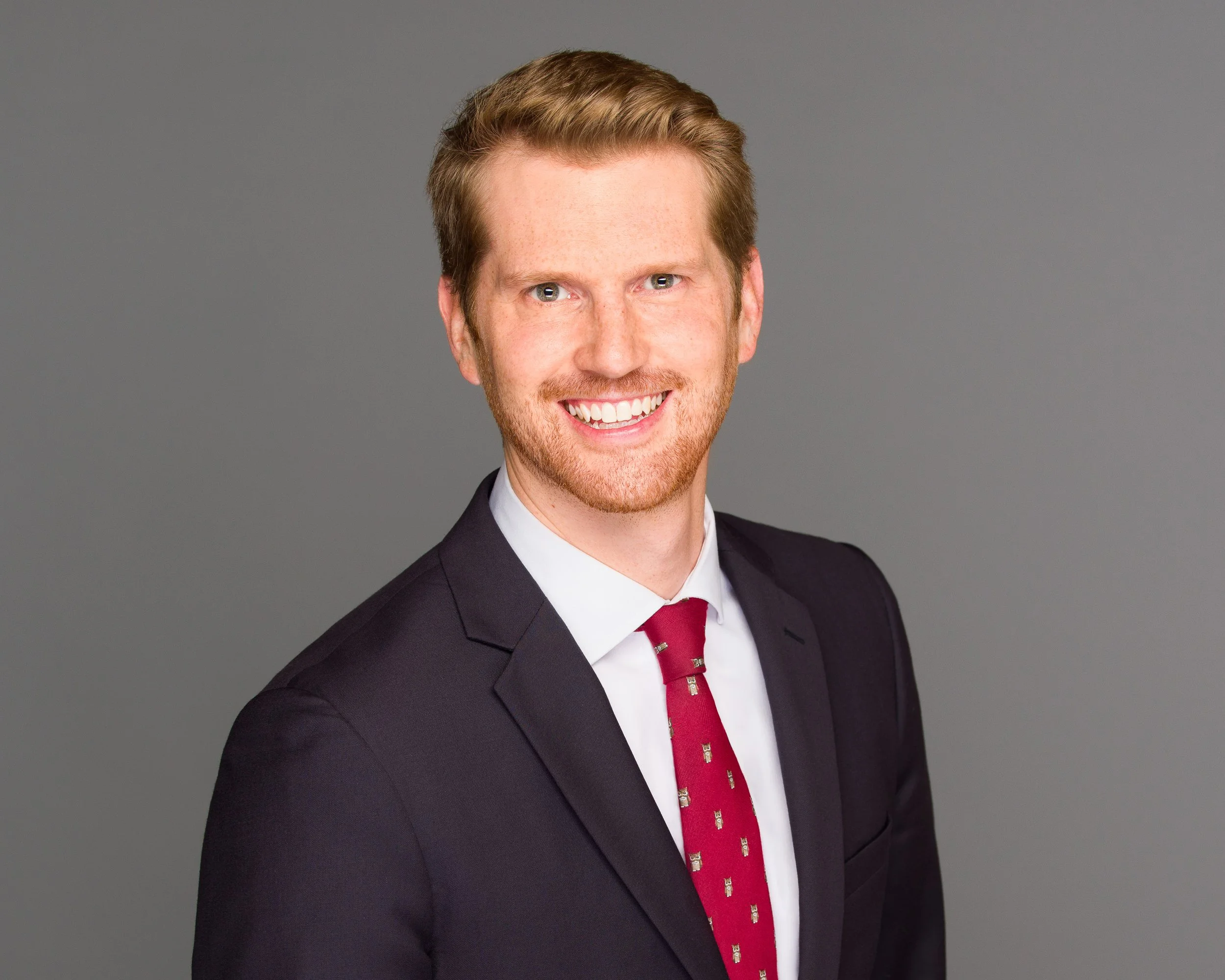 A smiling man with reddish hair and a beard, wearing a black suit, white shirt, and red tie, against a gray background.