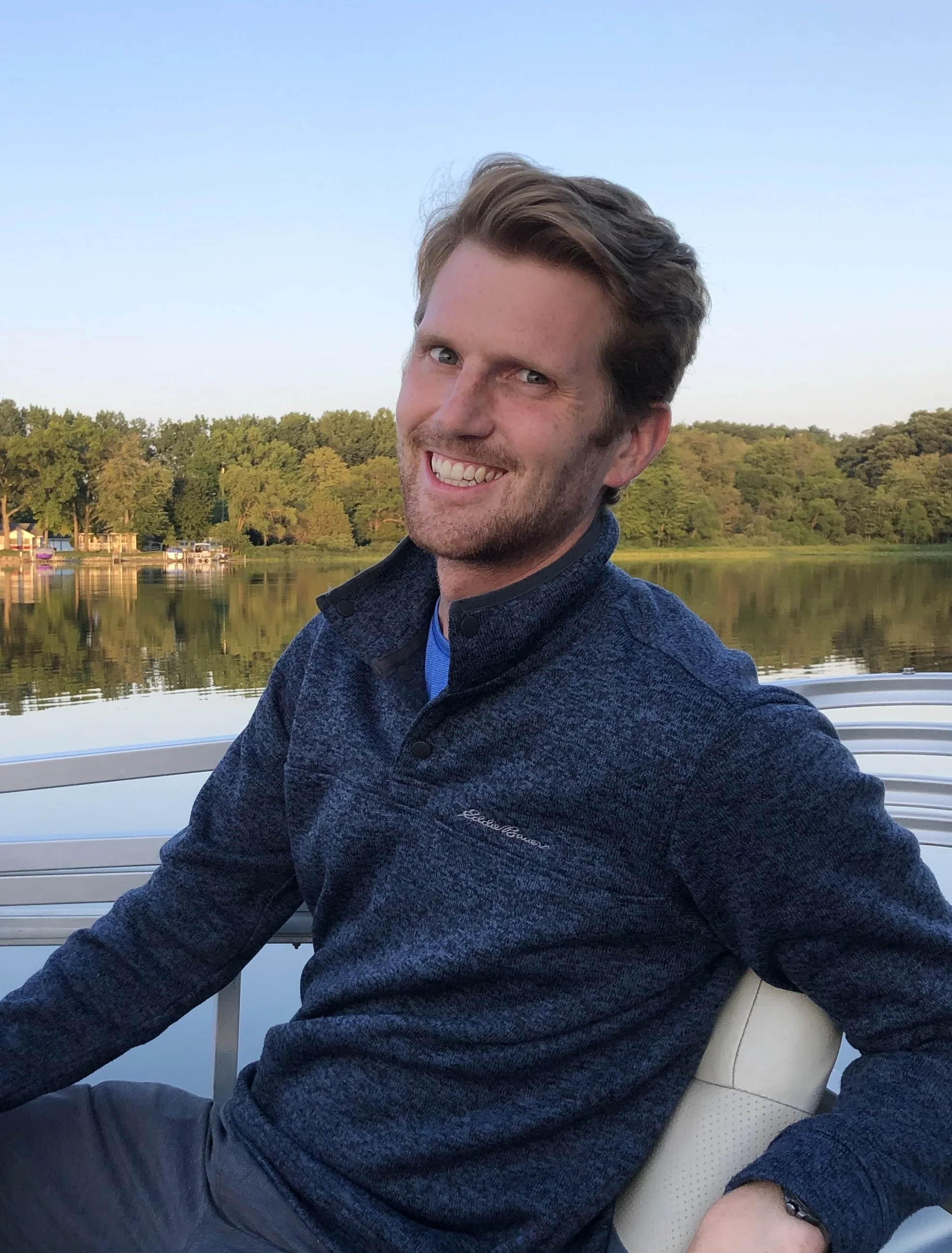 A smiling man with brown hair and blue eyes sitting on a boat with water and trees in the background during daytime.