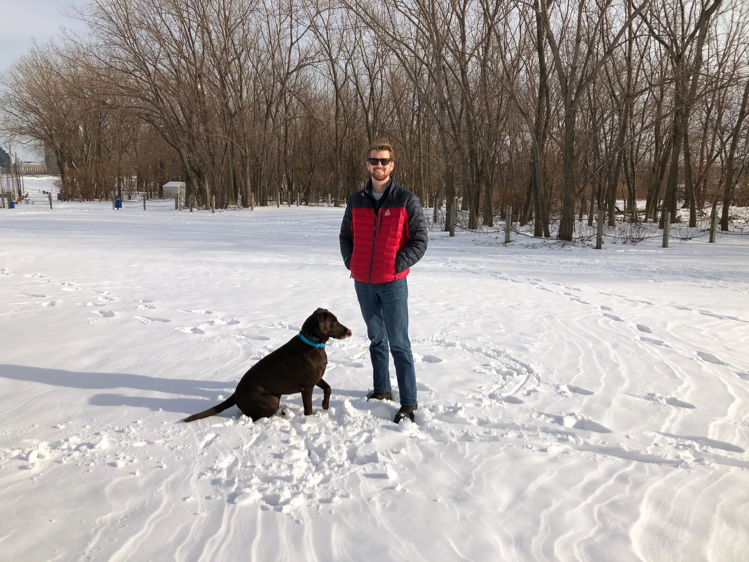 A man wearing sunglasses and a red and black jacket standing outside in the snow with a black dog sitting beside him, and a wooded area in the background.