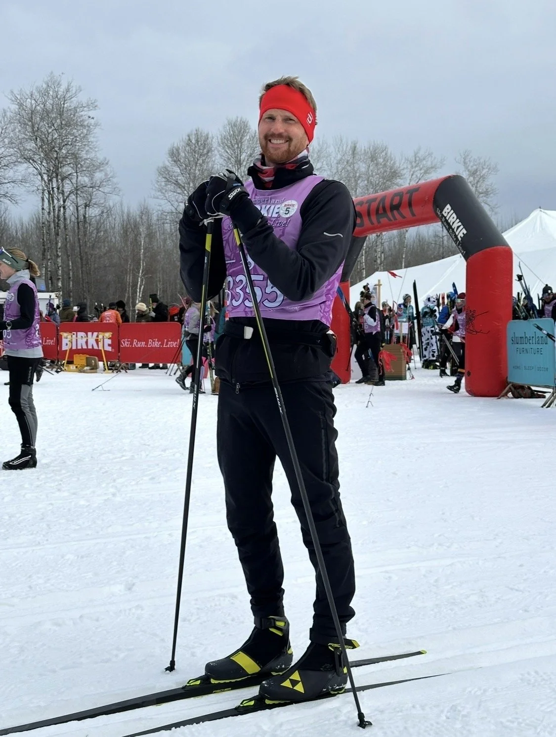 A man in winter sports gear standing on skis and holding ski poles at a snowy outdoor event, with a start arch and other participants in the background.