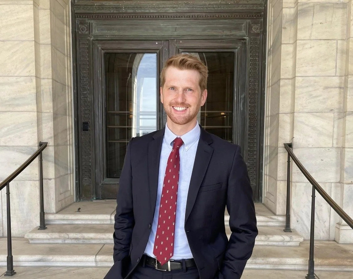 A man in a suit standing in front of a building with stone steps and an elaborate black door, smiling at the camera.