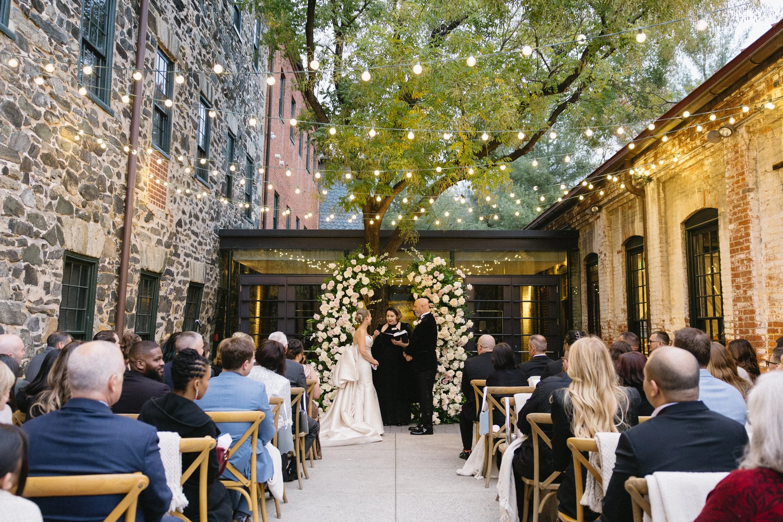 Wedding ceremony taking place outdoors under string lights, with a bride and groom exchanging vows in front of a floral arch, surrounded by seated guests in an urban courtyard with brick and stone buildings.