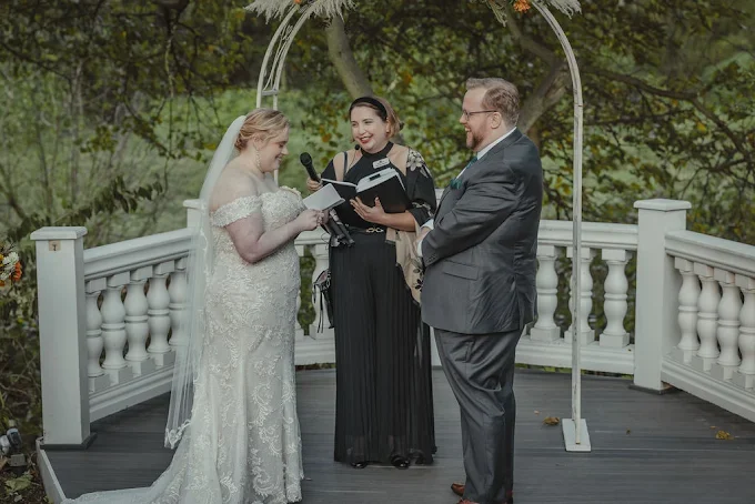 Bride and groom exchanging vows at outdoor wedding ceremony with officiant reading from book.
