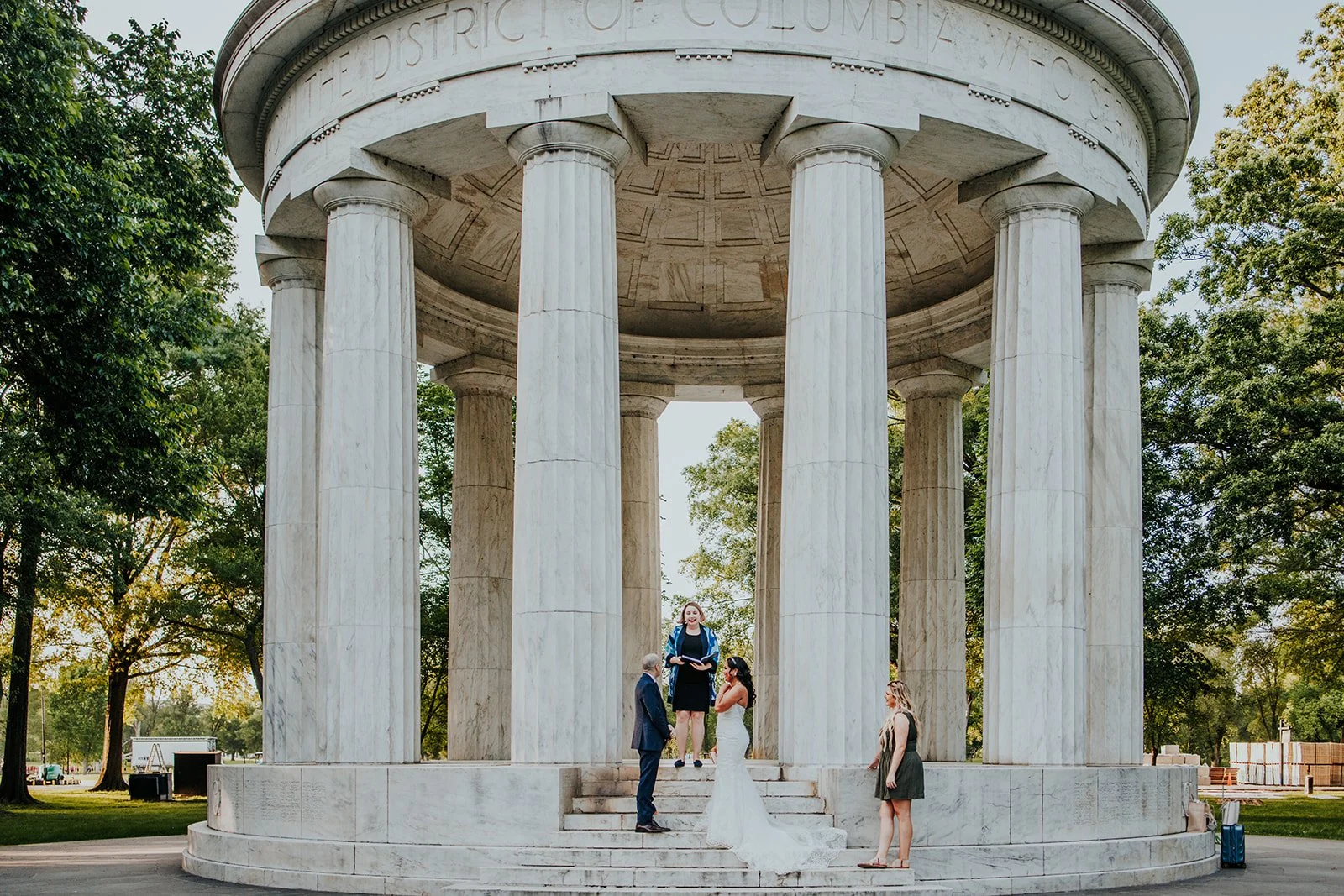 A wedding ceremony taking place under a large circular marble pavilion with columns, set in a park with trees.