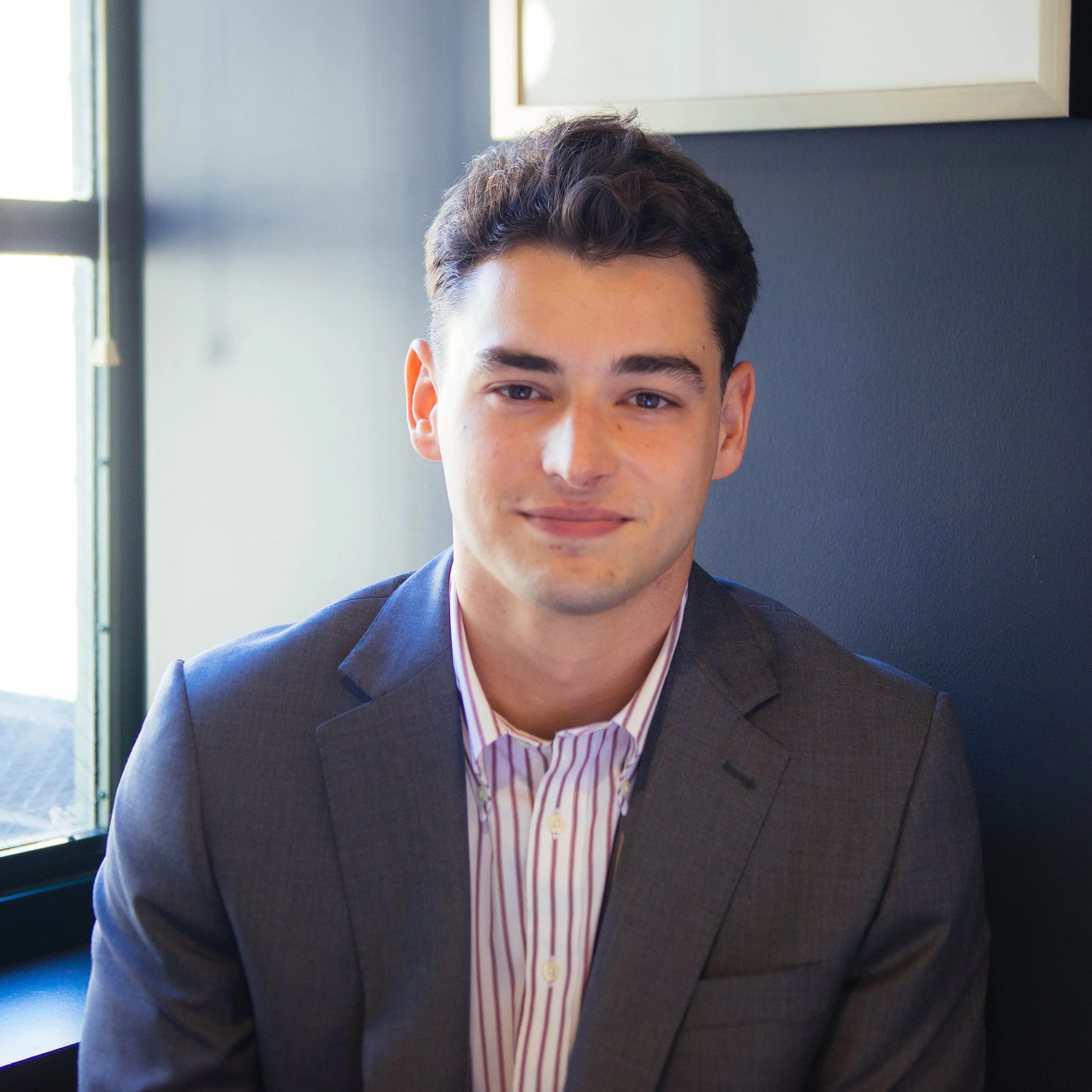 Close-up of a young man in a suit and striped shirt, indoors near a window with natural light.