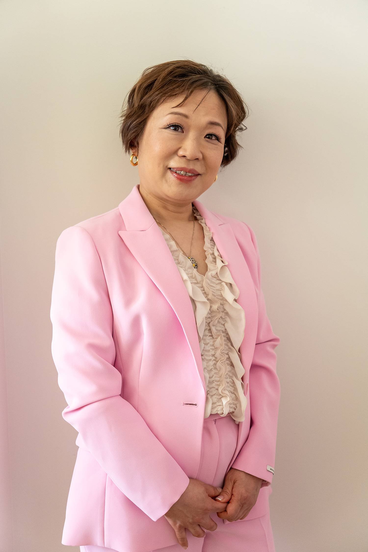A middle-aged woman with short brown hair smiling wearing a pink suit jacket and matching pants, with a beige ruffled blouse underneath, standing against a plain light-colored wall.