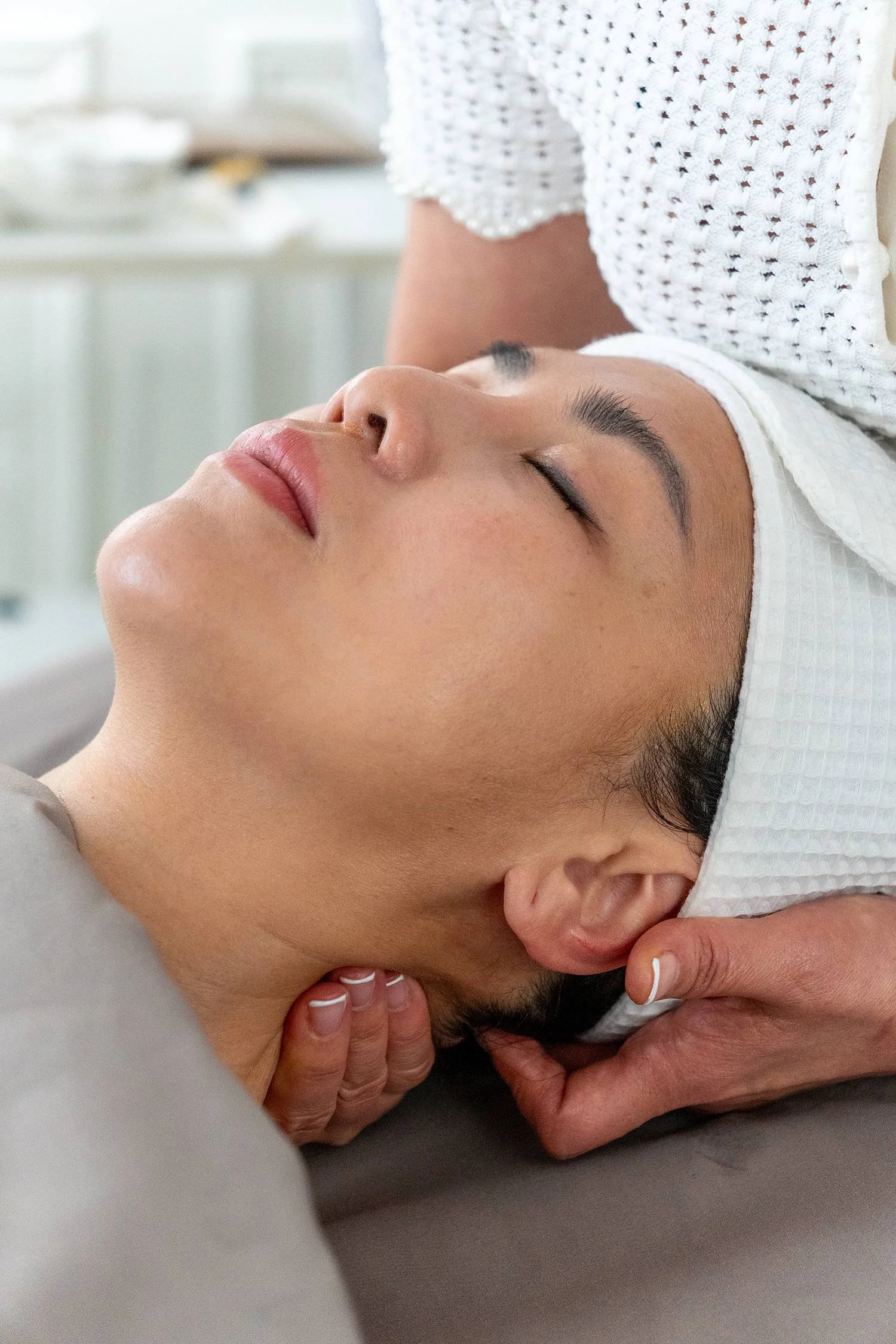 A woman with closed eyes receiving a facial massage in a spa or skincare clinic, lying on her back with a head band and a relaxed expression.