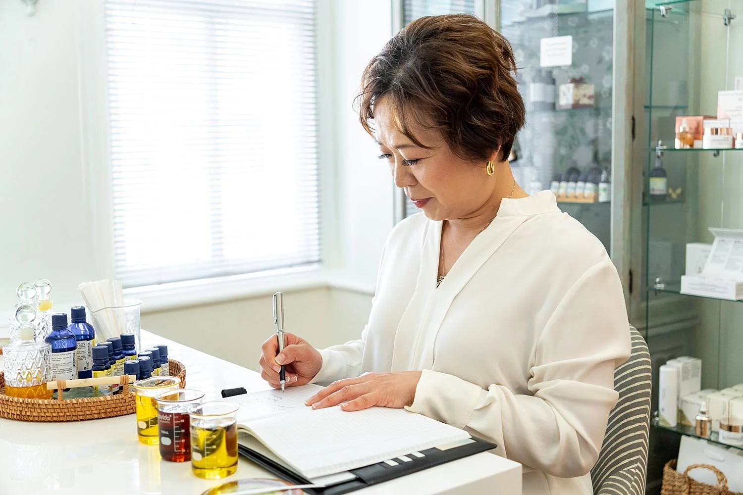 An elderly woman in a white coat writes in a notebook at a table with various skincare or essential oils and bottles, in a well-lit room with display shelves in the background.