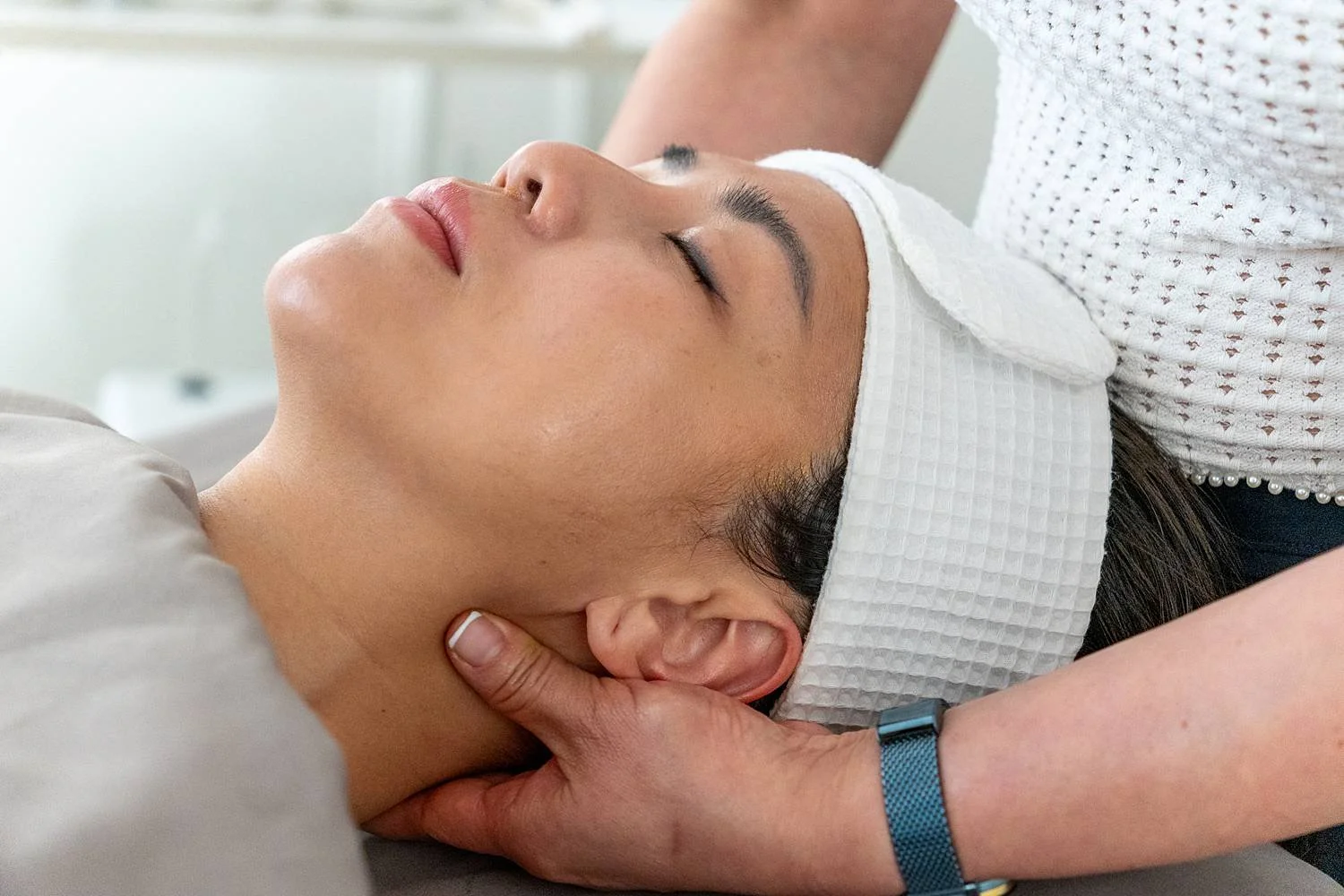 A woman receiving a massage with her eyes closed, lying on her back with a white towel headband during a spa treatment.
