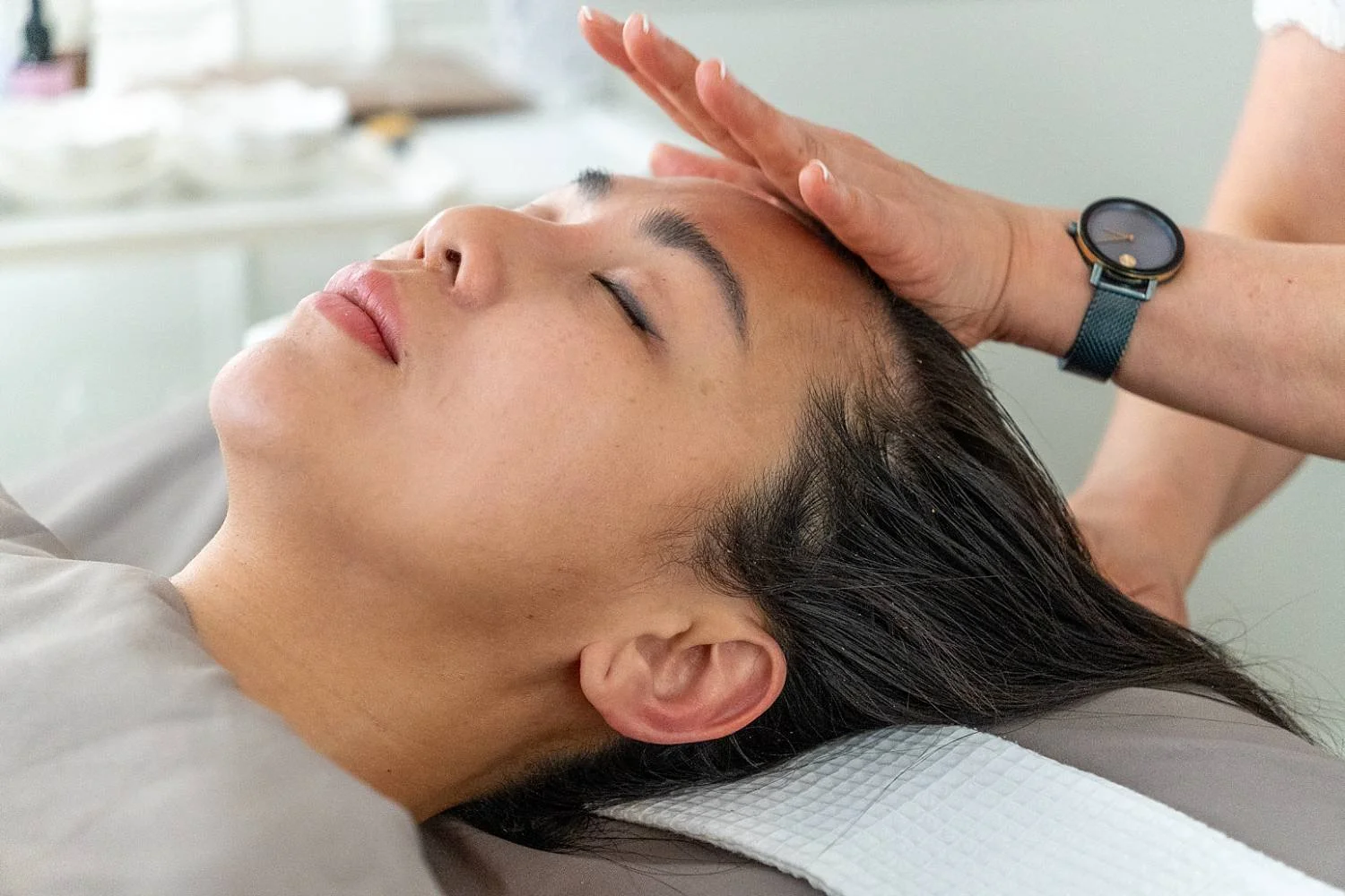 A woman receiving a facial massage from a therapist with a watch, lying on a treatment bed with her eyes closed