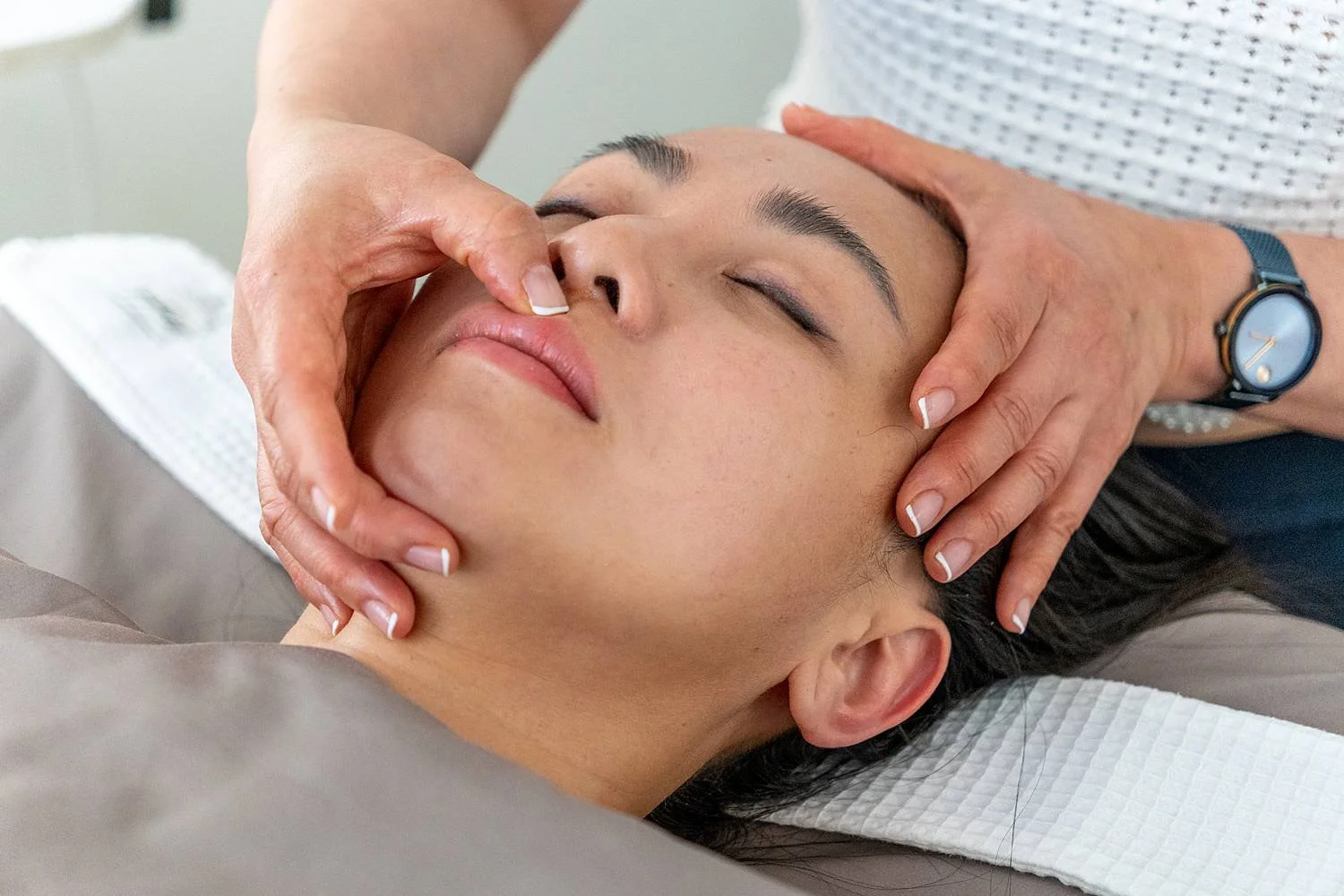 A person receiving a facial massage or skin treatment, lying down with eyes closed, while a practitioner gently massages the face and neck.