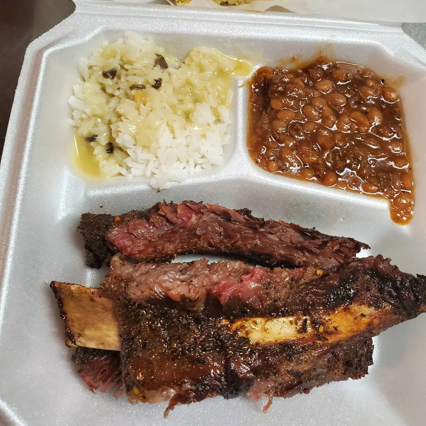 Foam container with grilled meat, rice with black beans, mashed potatoes with gravy, and baked beans.