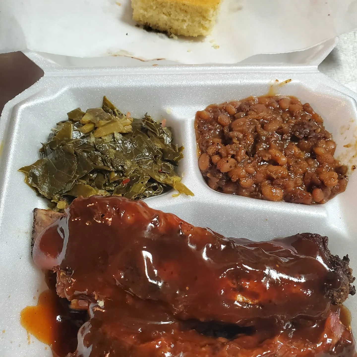 A styrofoam tray with collard greens, baked beans, and barbecue ribs with sauce.