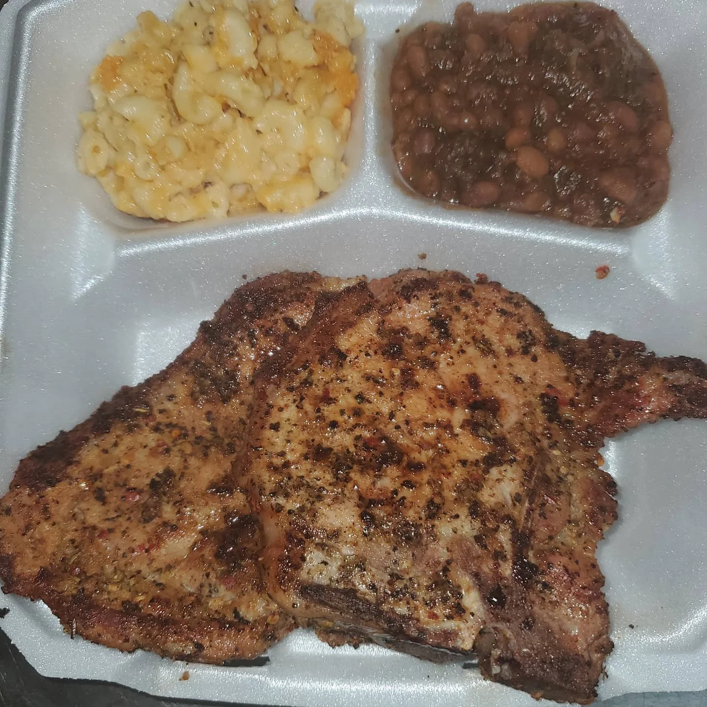 Fried chicken thigh with seasoning, macaroni and cheese, and baked beans on a divided white styrofoam plate.