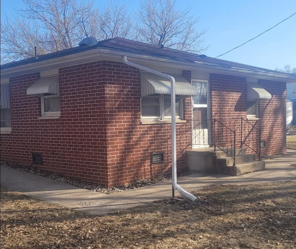 A small brick house with a metal railing front porch, three windows with striped awnings, and a white gutter downspout outside. There are leafless trees in the background and a clear sky.