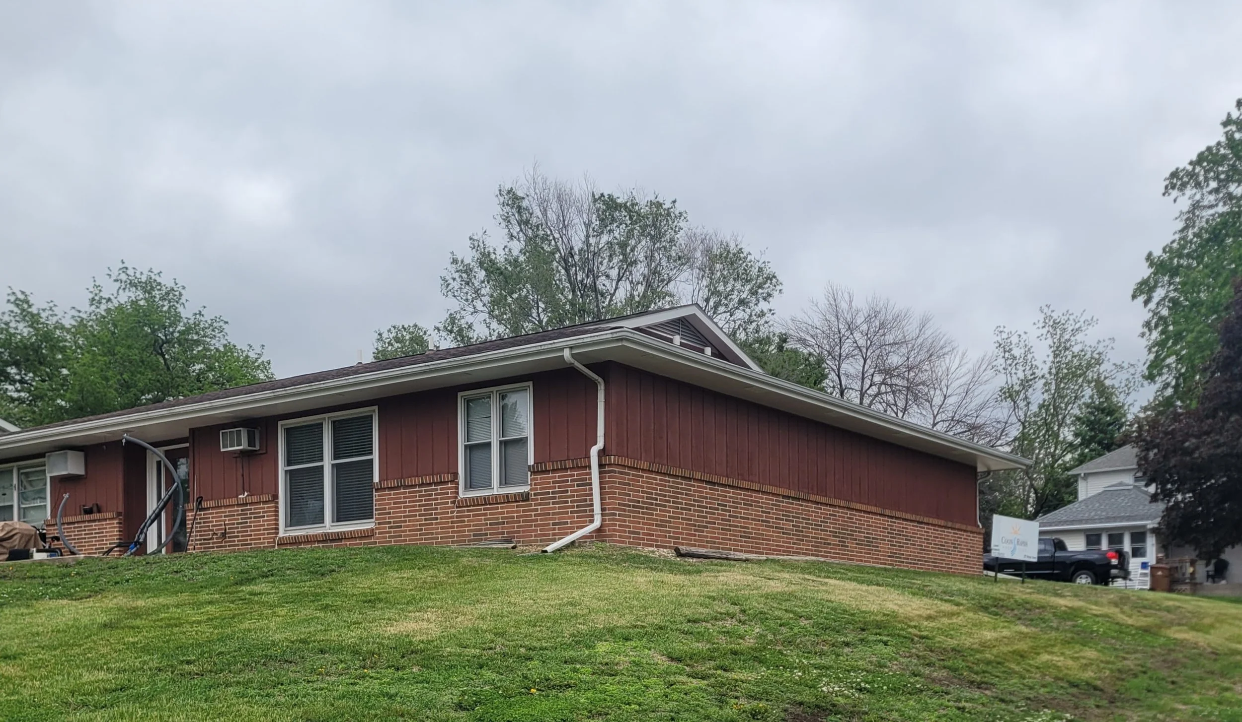 Front view of a two-story brick house with red siding on the upper part, white-framed windows, and roof with gutter system, surrounded by trees and a grassy yard under an overcast sky.