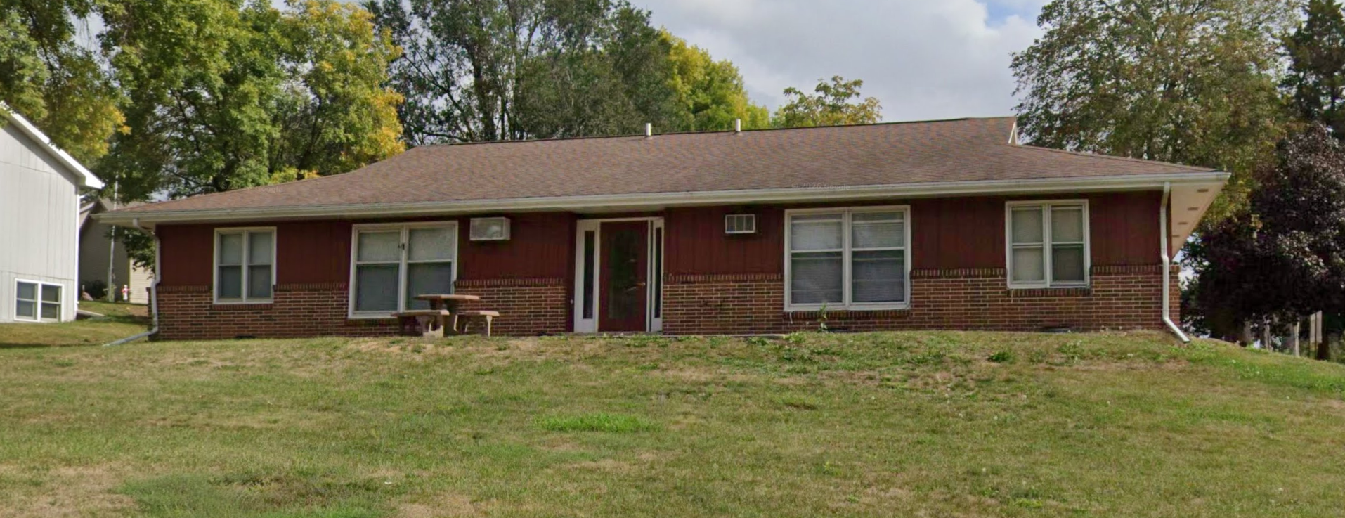 A single-story brick and wood house on a grassy hill, with four windows, a front door, a small outdoor table with benches, and trees in the background.