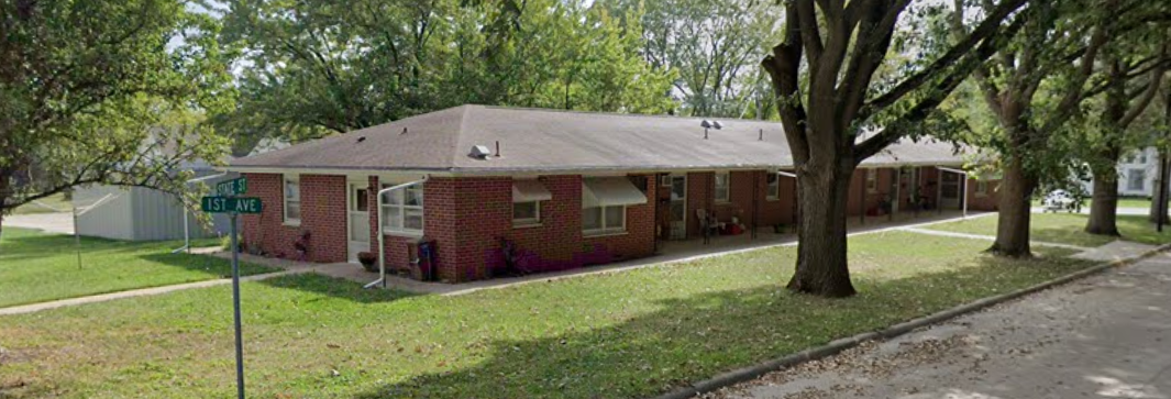 A single-story brick apartment building with a sidewalk in front, surrounded by green grass and large trees, located at the corner of 1st Avenue and State Street.